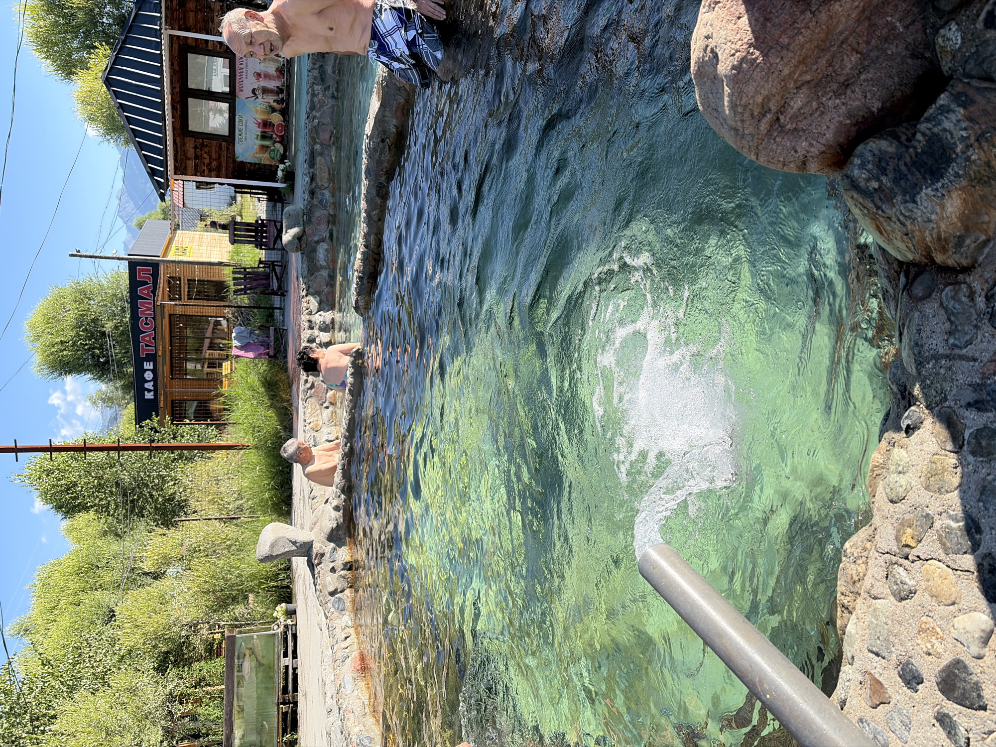 This media file shows an outdoor hot spring with several people enjoying the water. The water is a clear, greenish hue and appears to be well-lit by sunlight, suggesting a daytime scene. The setting includes natural rock formations around the edges of the spring and some vegetation and buildings in the background.
In the foreground, a pipe is visible, from which a stream of water is being actively dispensed into the hot spring, creating a visible disturbance on the water's surface. The people in the hot spring are partially submerged, some with their torsos visible. One individual, an older man, is seen from the chest up, looking towards the camera. Another person, wearing plaid shorts, is also partially visible. There are other individuals in the water, though less clearly depicted.
In the background, there are wooden buildings with signs. One sign, in Cyrillic script, reads "КАФЕ ТАСМАЛ" which translates to "CAFE TASMAL". Another sign above it appears to be advertising drinks. The buildings have a rustic, log-cabin style. The overall impression is one of a relaxed, recreational outdoor environment, likely a tourist spot given the presence of a cafe. The weather appears to be sunny and warm, contributing to the suitability for an outdoor hot spring.