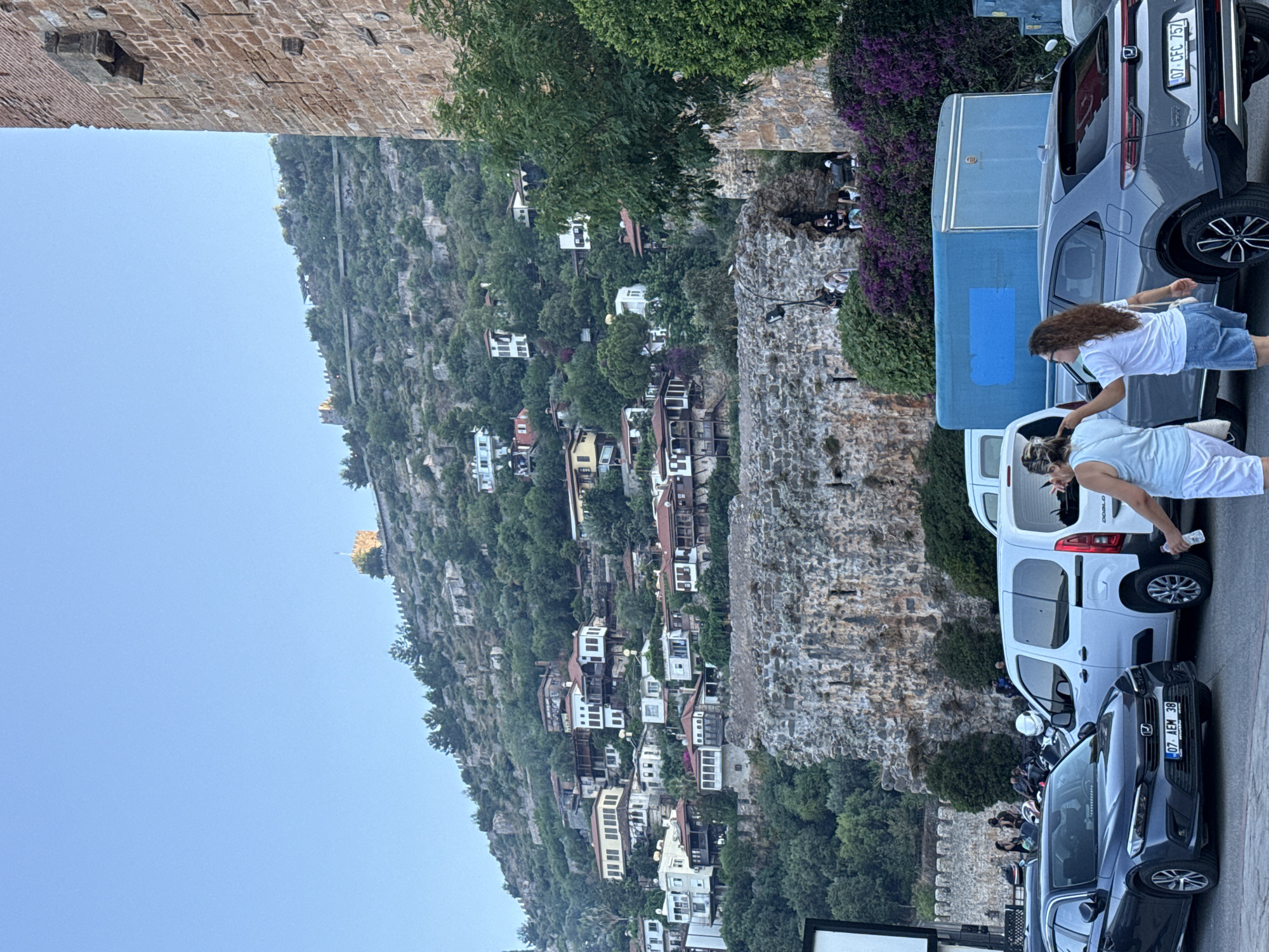 The image depicts a scene in Alanya, Turkey, showcasing the historic Alanya Castle perched atop a verdant hill dotted with traditional white houses with red roofs. In the foreground, several cars are parked, including a dark gray sedan with a Turkish license plate and a white van. Two women are visible, one wearing a light blue tank top and white shorts, and the other with curly brown hair, a white t-shirt, and denim shorts. They appear to be interacting, with the woman in the white t-shirt pointing. The backdrop features the ancient stone walls of the castle and lush greenery, with purple flowers blooming in the lower right section. The sky is clear and blue.