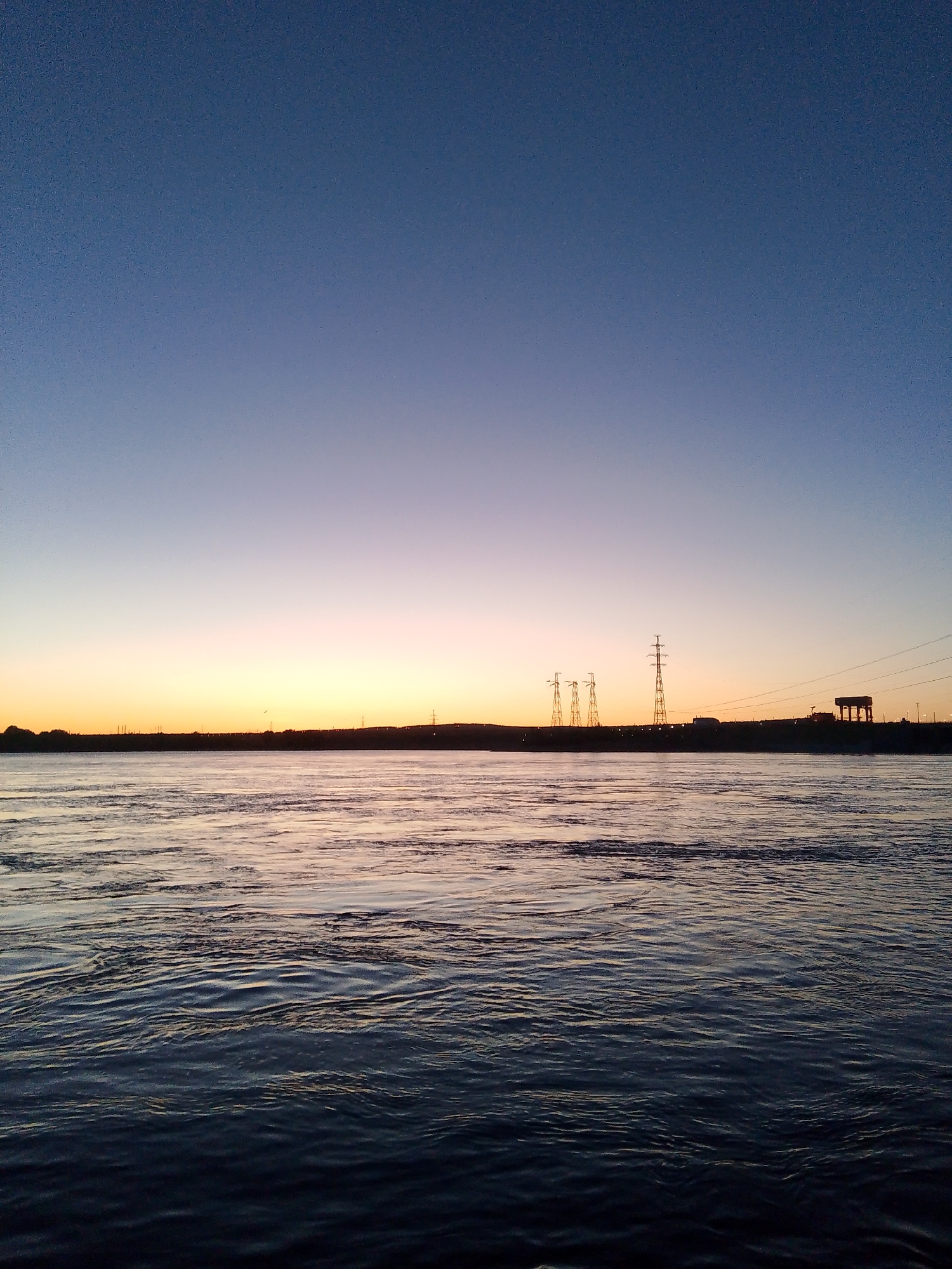 The image depicts a body of water, likely a river or large lake, under a gradient sky transitioning from a deep blue at the top to orange near the horizon. The water surface is textured with ripples and waves. In the distance, the dark silhouette of a landscape includes electrical transmission towers and some structures, possibly a power plant or industrial facility. The location is the Kama River in the city of Chaikovsky, Russia.