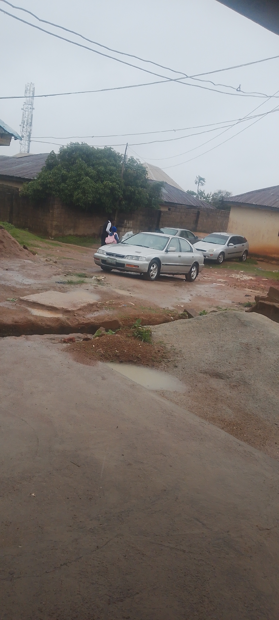 In a dusty, overcast environment, a silver sedan is parked on a dirt road. The car has its headlights on. Two individuals, one carrying a pink backpack, are standing near the vehicle. The background shows a brick wall, trees, and a telecommunications tower. The ground is uneven and appears to be wet in places, with puddles reflecting the sky. Piles of dirt or gravel are visible in the foreground.