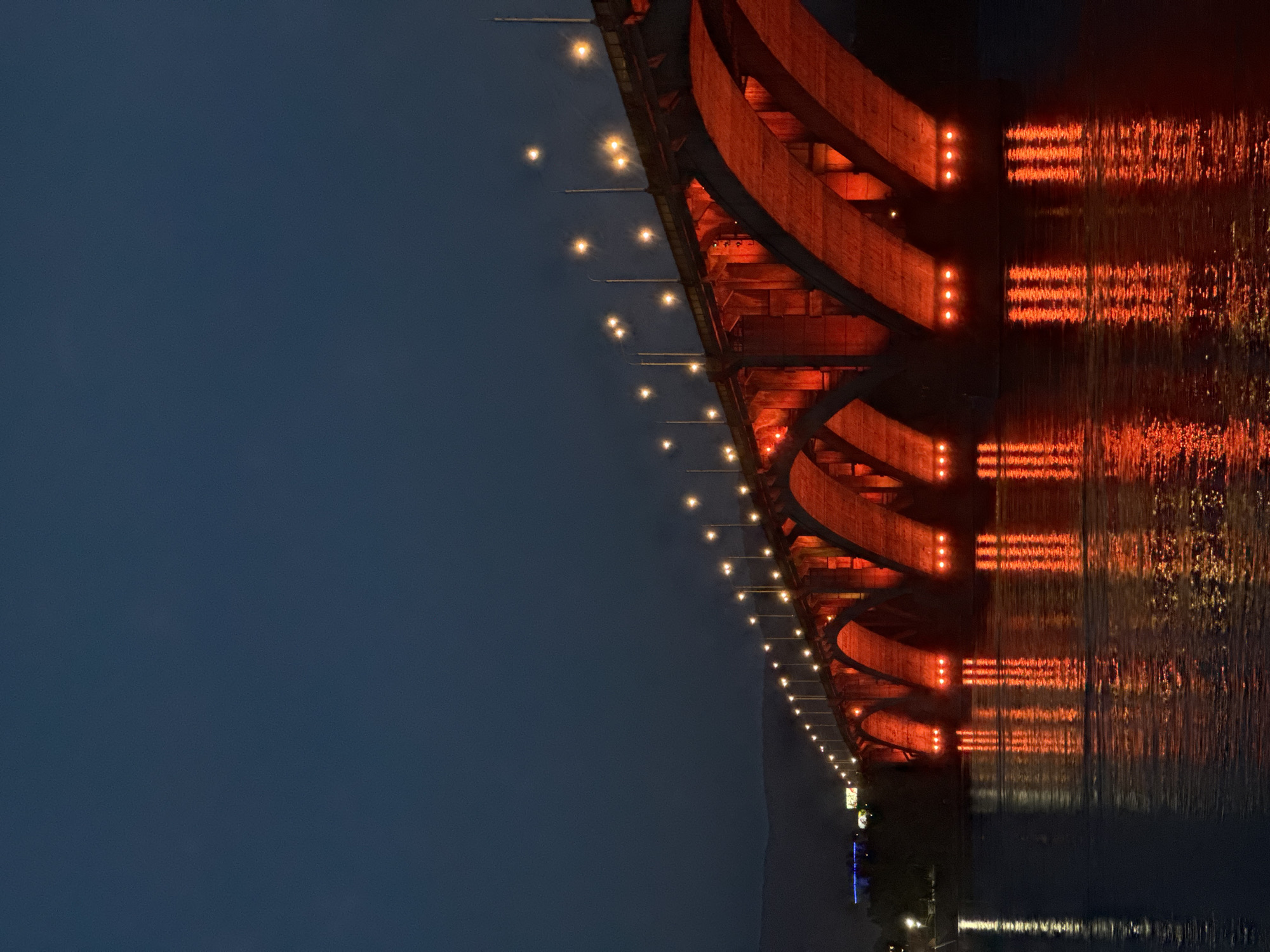 The image shows the Kommunalny Bridge in Krasnoyarsk, Russia, at night. The bridge is illuminated with red lights, which are reflected in the Yenisei River below. The sky is dark and cloudy, and there are no people visible in the image. The bridge is made of concrete and has a series of arches. The lights on the bridge are evenly spaced and cast a warm glow on the water. The reflection of the bridge on the water is distorted by the ripples, creating a beautiful and dynamic effect. The overall mood of the image is peaceful and serene, with a touch of mystery due to the low light conditions and the striking red illumination.