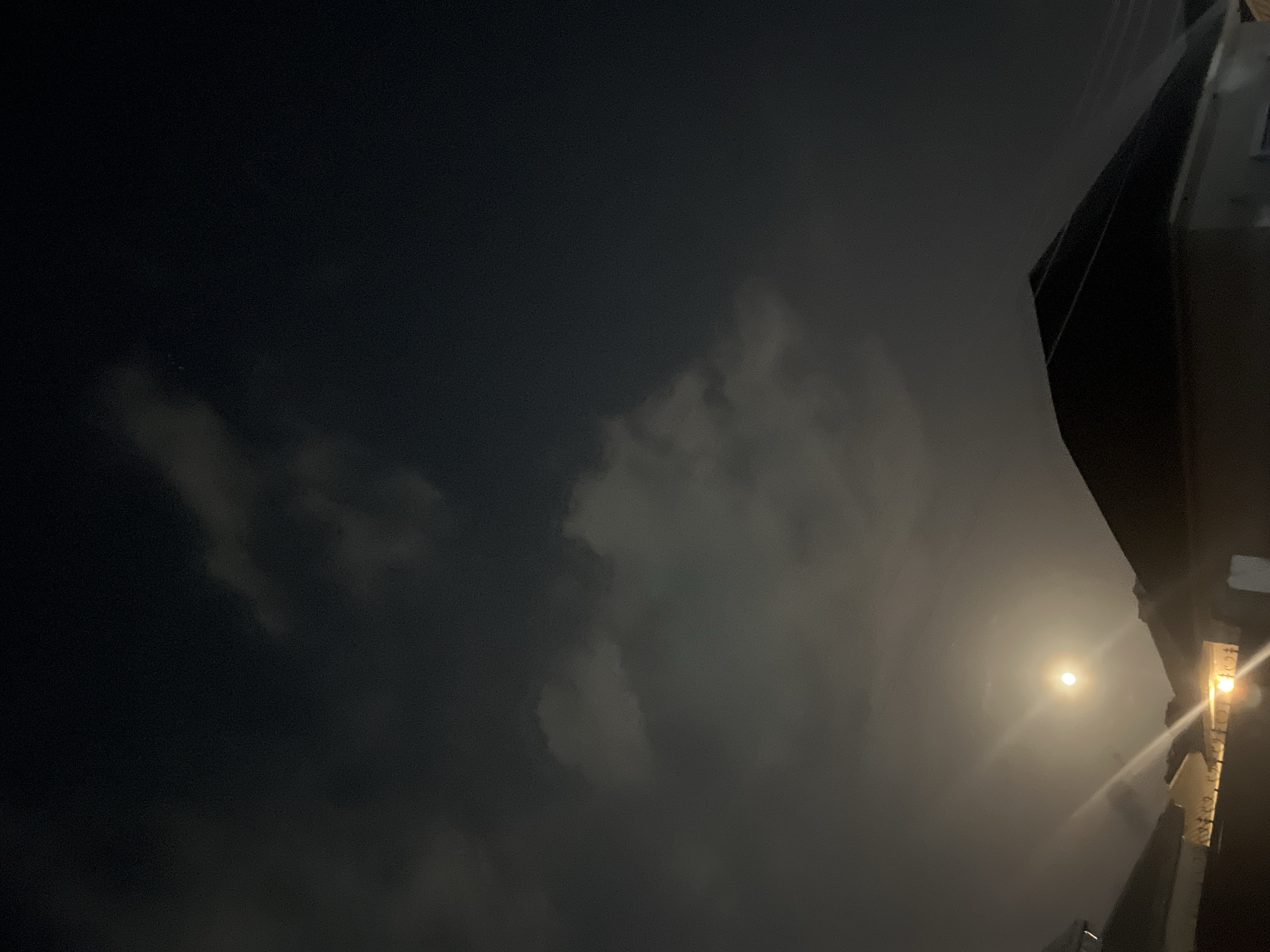 A nighttime scene in Abuja, Nigeria, shows a dark sky with scattered clouds. A bright light source, likely the moon or a streetlamp, illuminates the clouds and casts a glow in the lower-right portion of the frame. The silhouette of a building or structure is visible in the upper-right, and what appears to be a streetlight or illuminated sign is also present on the right edge of the image. No people are visible.