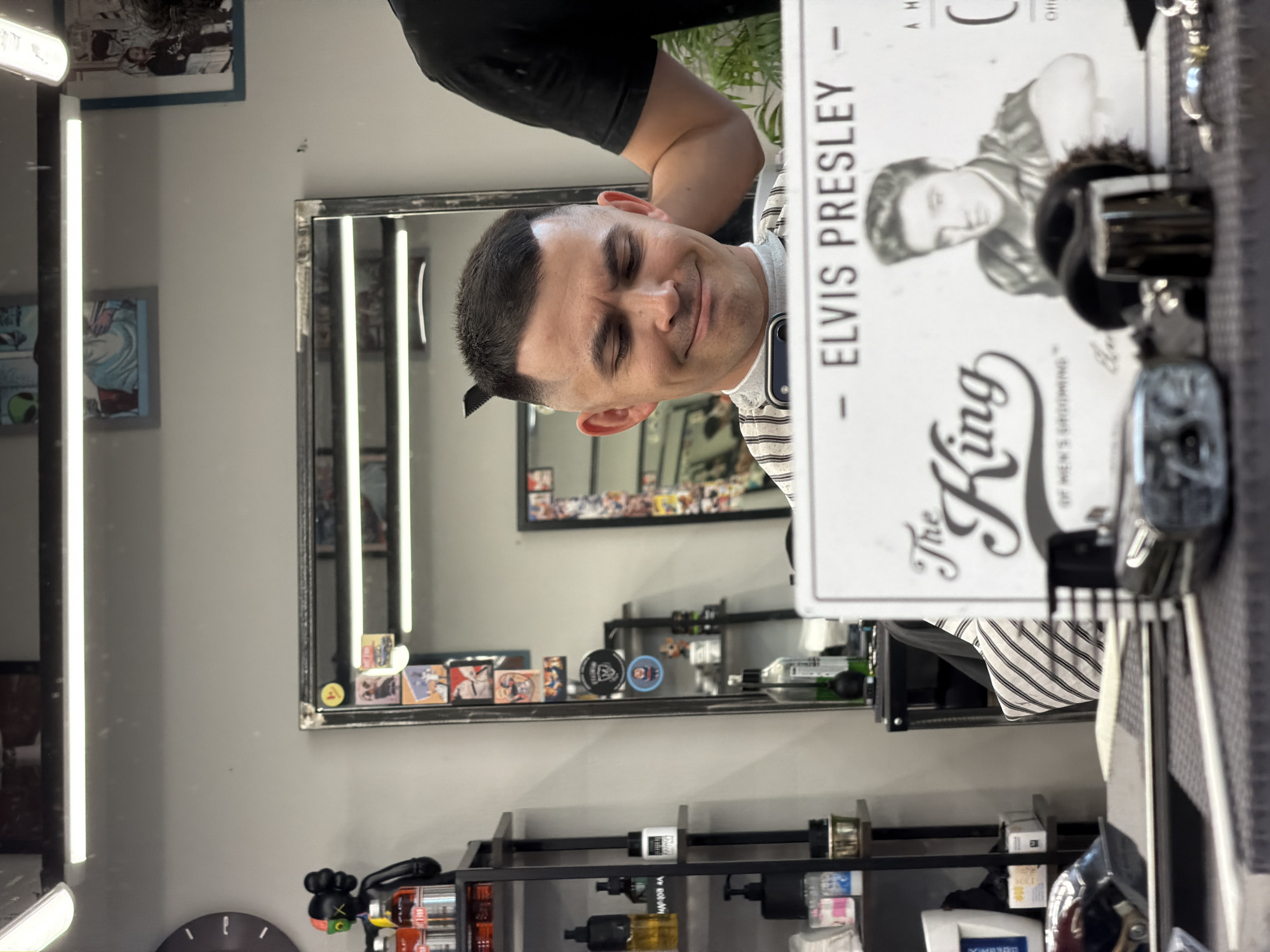 A photograph shows a man with a fresh haircut in a barbershop. He is wearing a striped shirt, and his hair is short and neatly styled. A comb is tucked into his hair near his ear. The barber is visible in the background. A mirror behind the man shows the barber's equipment and products on shelves. In the foreground, a sign with an image of Elvis Presley and the words "The King of Men's Grooming" is partly obscuring the view. The barbershop appears to be well-lit with modern light fixtures. The location in Izhevsk, Russia, is not possible to determine from the image.