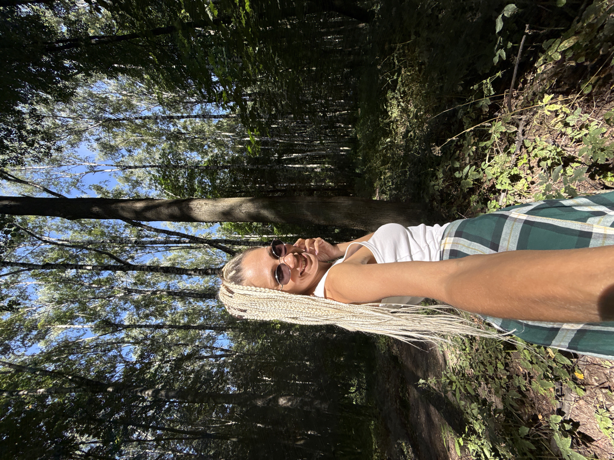 A woman with long blonde braids and sunglasses is taking a selfie in a forest. She is wearing a white tank top and a green and white plaid skirt. The forest has tall trees and lush green foliage. The sun is shining through the trees, creating dappled light on the ground. The location is identified as Rusanovo, Russia.