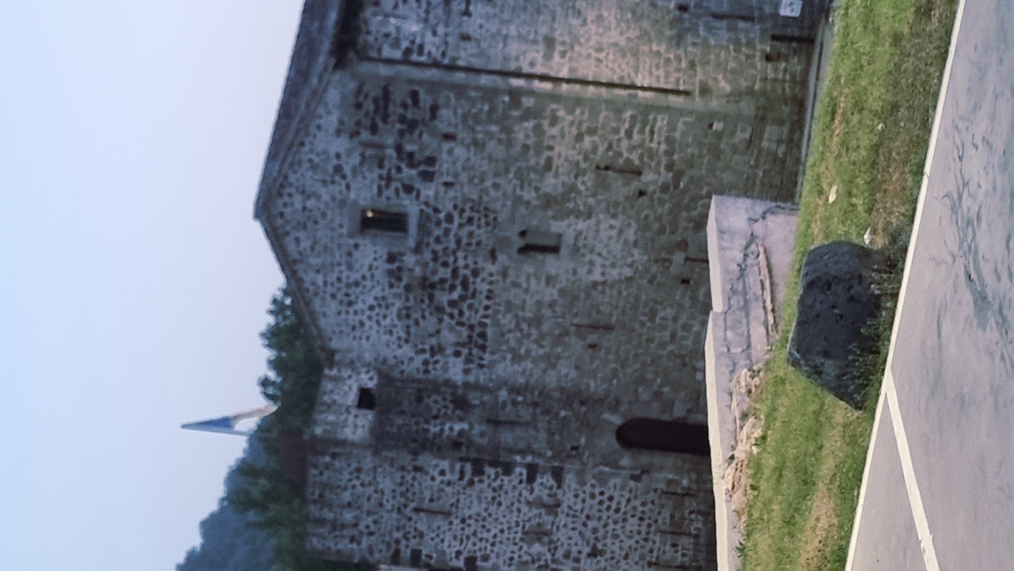 The image captures a view of a stone building with a pitched roof, possibly a church or historical structure, situated in Sant Joan Les Fonts, Spain. A flag, appearing to be tri-colored with blue, white, and possibly red or black stripes, is visible flying in the distance above a tree line and a hilly landscape. In the foreground, a patch of grass is visible with a large, dark rock placed upon it. A paved surface with white markings is also present in the lower portion of the frame. The sky is a pale, overcast hue, suggesting it might be dawn or dusk. The stone building features small, dark windows and arched doorways, indicating its age and architectural style. The overall scene is one of historical architecture and natural landscape under a muted sky.