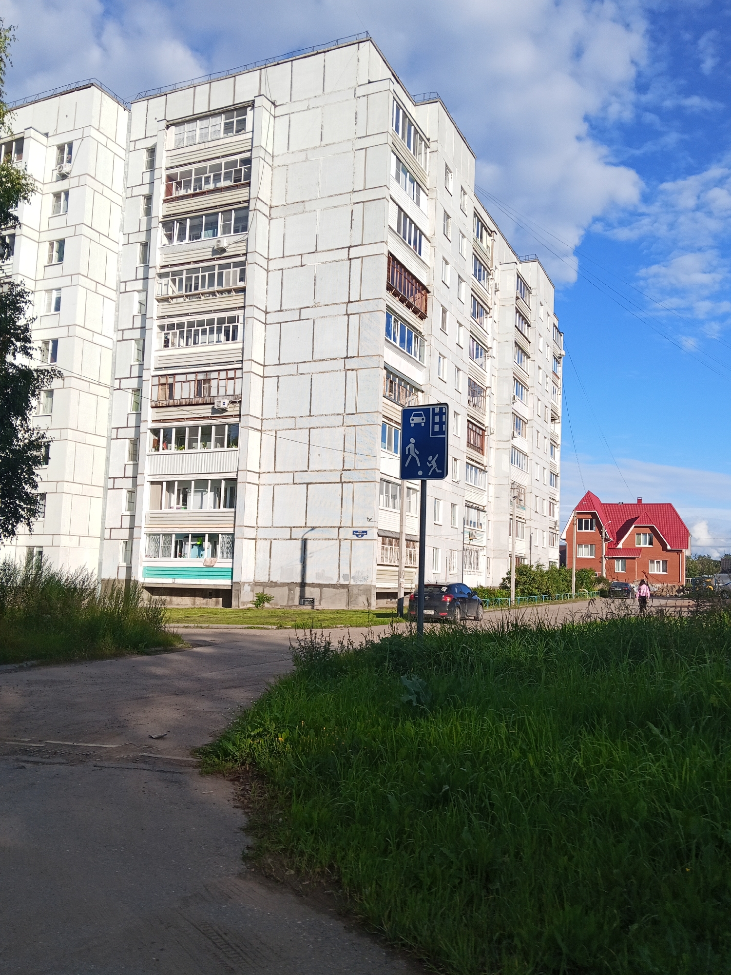 The image depicts a residential area in Chaikovsky, Russia, characterized by a large, multi-story apartment building and a smaller, detached house. The apartment building, painted in a light gray color, features numerous balconies and windows, with some air conditioning units visible. A blue traffic sign with pedestrian and car symbols is positioned near the apartment building. A black car is parked on the street in front of the building, and another dark-colored car is further down the road. A person, possibly female, wearing pink clothing, is walking on the sidewalk. The house is a brick structure with a prominent red roof. The foreground is dominated by lush green grass, and a paved road or pathway is visible in the lower portion of the image. The sky is blue with scattered white clouds. The scene appears to be set during daylight hours.