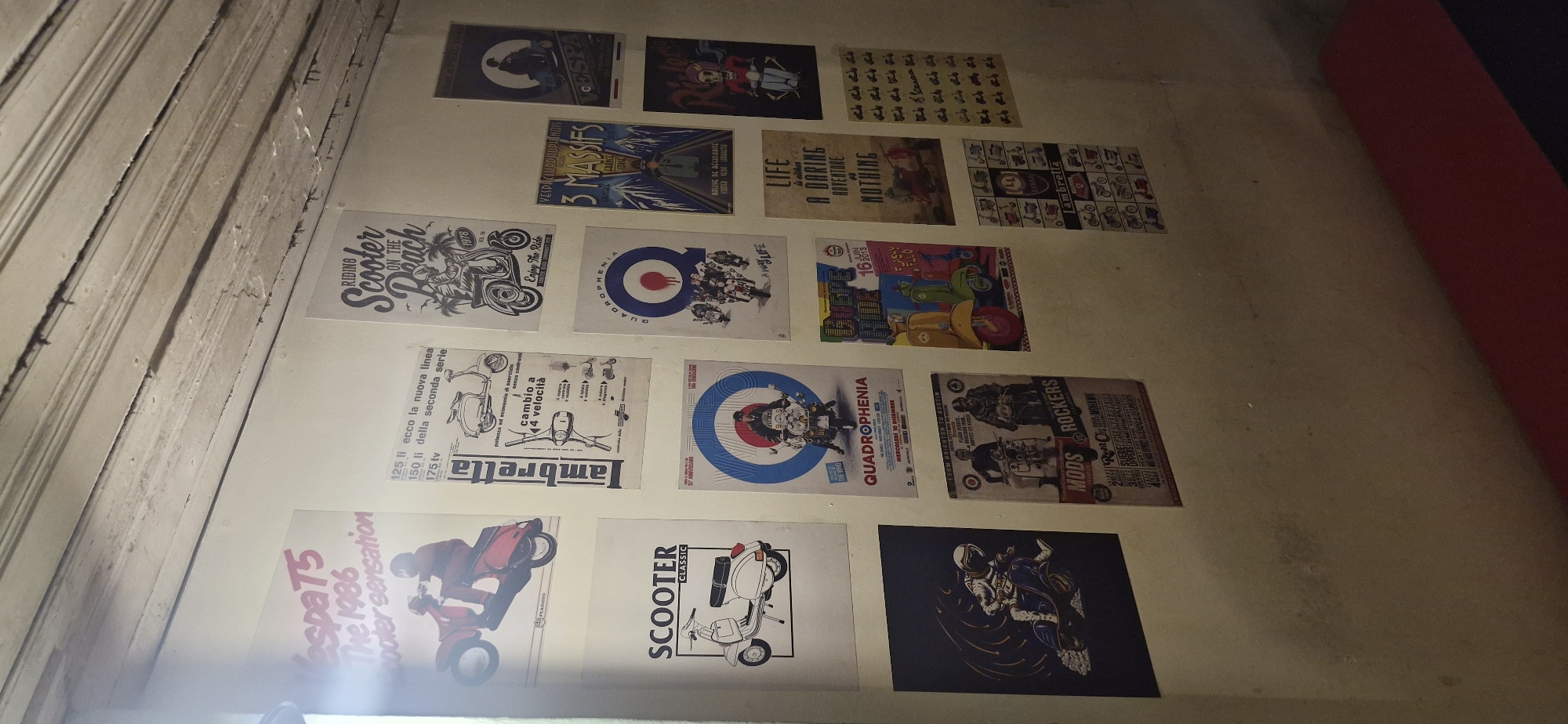 The image shows a wall in what appears to be an indoor space, possibly a room or a shop, decorated with numerous posters. The wall is a pale, muted color and has a textured appearance, with some visible imperfections and shadows. The ceiling above the wall is made of wooden planks, some of which appear worn and have dark lines or cracks.
The posters are predominantly featuring scooters, with a focus on vintage or classic models. They are arranged in a grid-like fashion, but with some overlap and variation in size and orientation. Some of the posters have text in Italian, suggesting a European influence or origin. The posters depict various scooter models, advertisements, and artwork related to scooter culture, such as "Quadrophenia," "Scooter Classic," and what appears to be a "Vespa T5." There are also posters with graphical designs and text that are not immediately decipherable without closer inspection.
On the bottom right of the image, a portion of a red upholstered surface is visible, which could be a bench or seating.
The image does not provide any specific visual cues to pinpoint its location within Rantau, Indonesia. The content of the posters, primarily related to European scooter culture, does not align with any known local context of Rantau.
