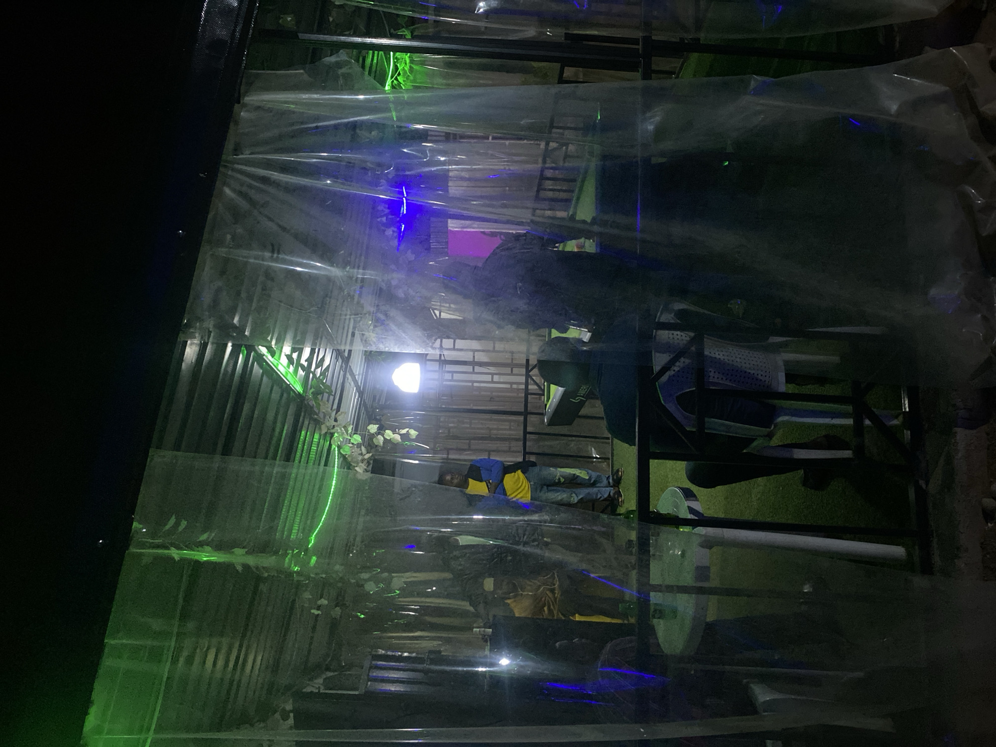The image appears to depict an outdoor setting, possibly a restaurant or entertainment venue in Jos, Nigeria, enclosed by a structure of metal framing and translucent plastic sheeting. Green and blue lights are visible, casting an eerie glow. Inside, a person is lying on what appears to be a platform or elevated surface. The interior contains tables, chairs, and other furnishings. The ground outside the structure is partly covered with artificial turf. The scene seems to be at night.