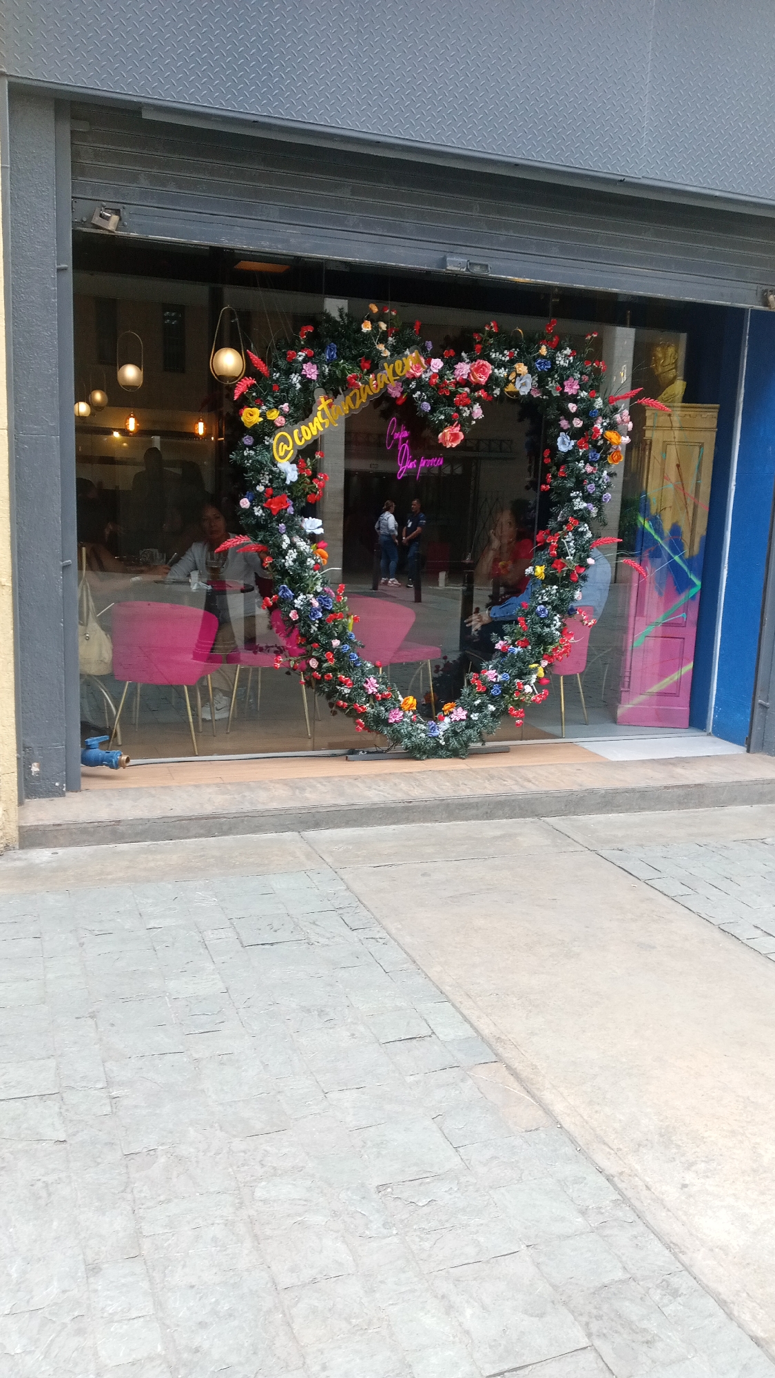 Here is a description of the image:
The image shows the storefront of a business, likely a cafe or restaurant, with a large floral heart-shaped decoration displayed in the window. The heart is made of various colorful flowers and greenery. Inside, patrons are seated at tables with pink chairs. There's a neon sign with the name of the place, "@constanzhakeem" above the floral heart and "Corpa Dos proeces" below. People are visible through the window, suggesting normal business activity. The exterior consists of a gray pavement. The store front window has a gray steel shutter door above the glass. The location, in the city of Unknown, Unknown, is not clear.