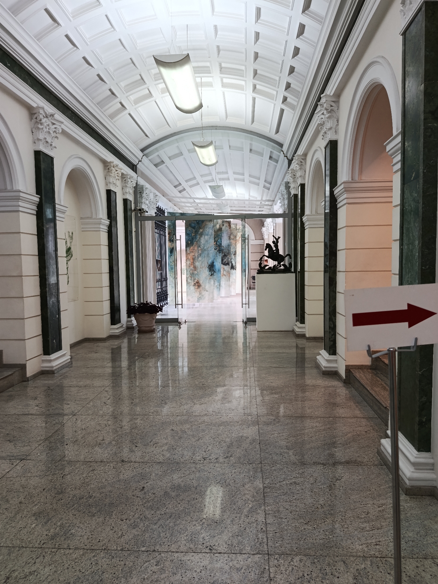 The image shows a long, well-lit interior hallway with a high, coffered ceiling and large rectangular fluorescent light fixtures. The hallway is lined with white columns and arches, with dark green marble accents. The floor is covered in large, polished granite tiles that reflect the light and the architectural details. In the foreground, on the right side, a white sign with a red arrow points to the right. Further down the hallway, through glass doors, a mural with abstract colorful patterns is visible. A black sculpture of a horse and rider stands on a white pedestal in the middle of the hallway. A potted plant sits in a terracotta pot near the left wall. The hallway appears to be empty of people.