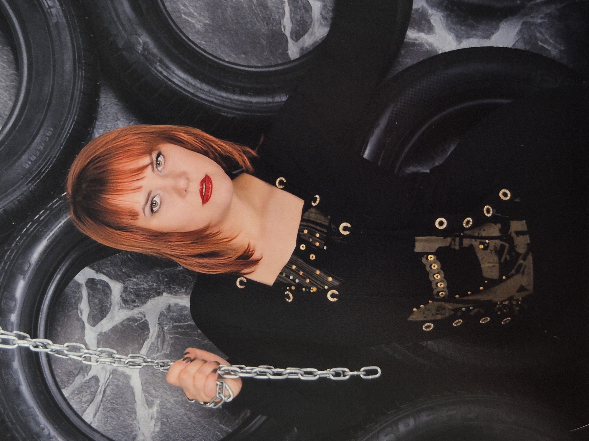 The image shows a young woman with red hair and red lipstick holding a metal chain. She is wearing a black top with gold accents. She is standing in front of a background of stacked tires. There is no indication of the location being Schwäbisch Gmünd, Germany, as the image provides no contextual information for it.