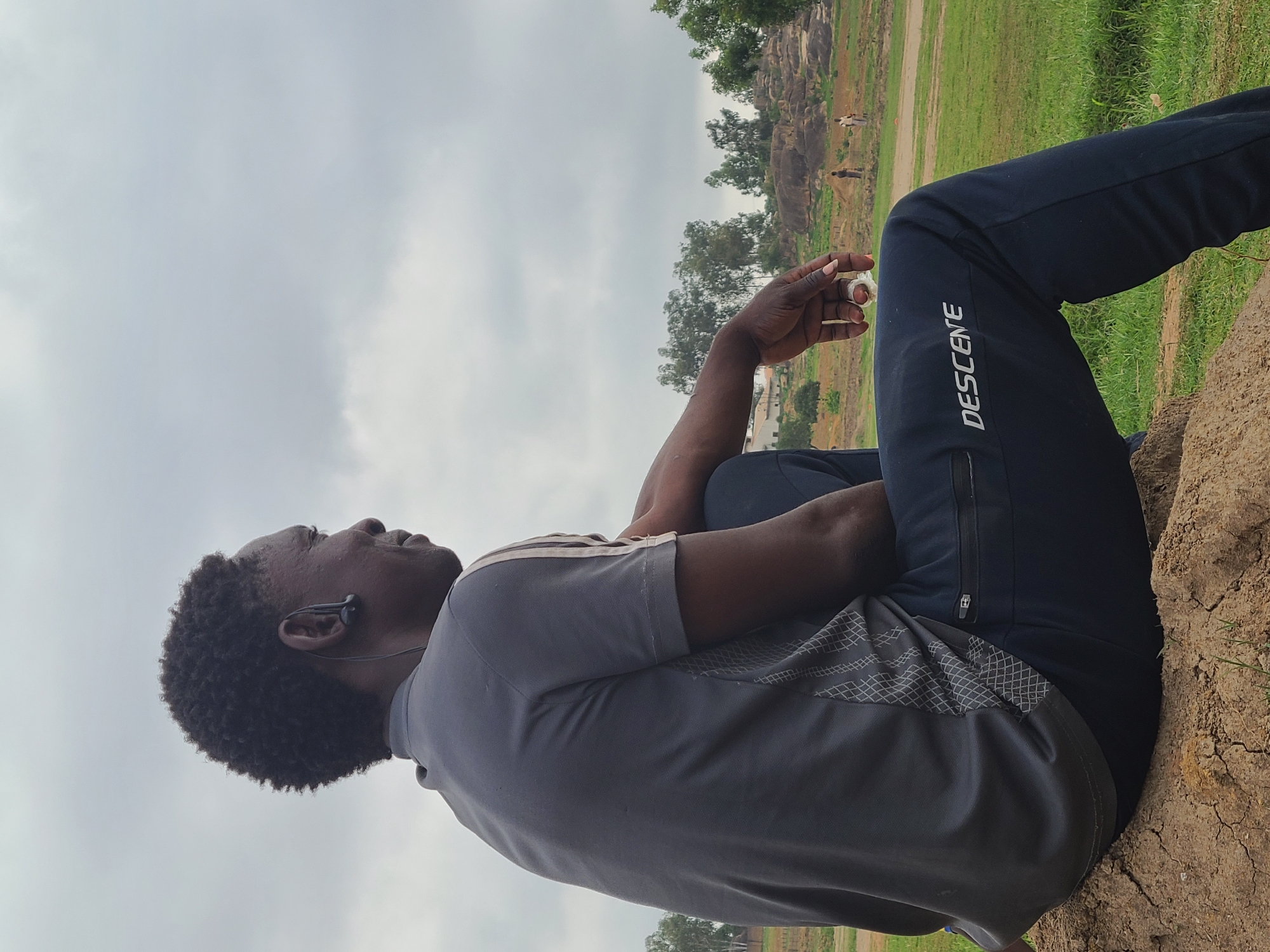 A man with dark skin and short, curly dark hair is depicted sitting outdoors, viewed from a low angle, looking upwards and slightly to his left. He wears a grey short-sleeved polo shirt with a patterned design on the lower right side and dark blue track pants with a white vertical "DESCENTE" logo visible on his left leg. A black wired earbud is inserted in his right ear. His right arm is bent, holding a small, crumpled white object, while his left arm is bent and rests across his body. He appears to be seated on a dry, cracked earth or rock surface.
The background features a natural landscape under an overcast, cloudy sky. Lush green grass and various trees are visible behind him. Further in the distance, rocky hills or formations can be seen, with what appears to be faint figures of other people or animals in the far background. The overall impression is one of calm contemplation or listening in an outdoor, possibly rural or semi-rural setting on an overcast day.
