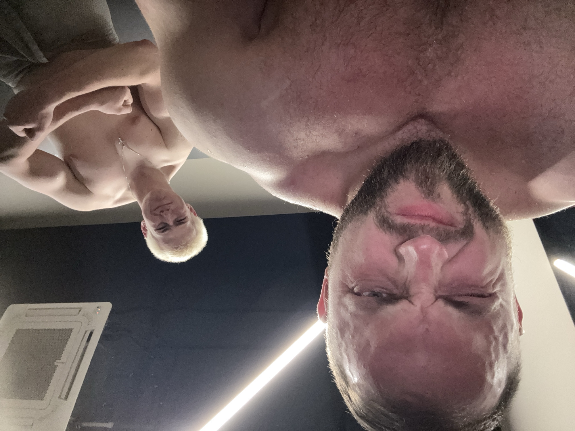 The image presents a low-angle, inverted view of two shirtless men indoors. The closest man, dominating the right side of the frame, has dark hair and a beard. His face is contorted in a grimace, with his eyes squeezed shut, and his skin appears sweaty.
Behind and to the left of him is another man with short, bright blonde hair and a clean-shaven face. He is looking directly at the camera with a slight smirk and wears a silver pendant necklace. His arms are bent, and his chest is prominent, suggesting a posed display of physique. His skin also appears glossy with sweat.
The setting is an interior space with a dark ceiling or wall, illuminated by artificial light sources. A long, bright linear light fixture is visible in the background, near the bearded man's head. In the bottom-left corner, a white square ceiling panel, possibly an air vent or a type of light, is partially visible. The overall scene suggests a workout environment or a similar context where individuals might be showcasing their bodies after physical exertion. No visible text is present in the image.