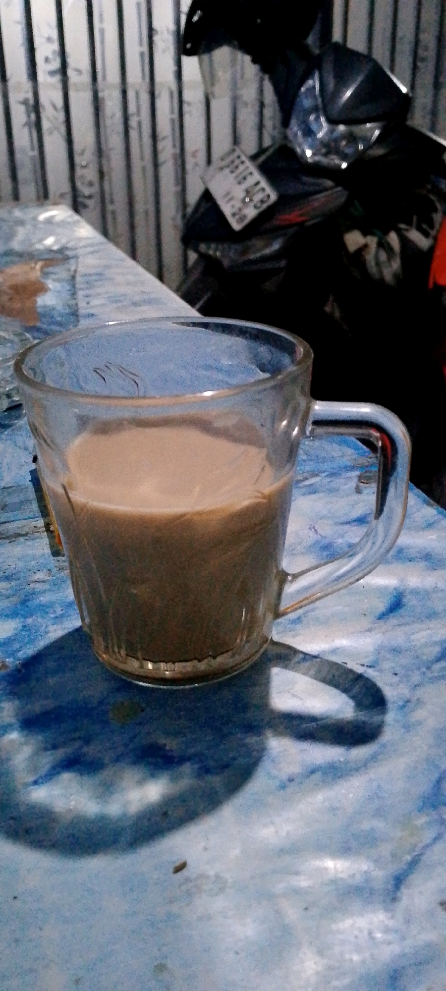 A clear glass mug filled with a light brown beverage, possibly coffee or tea with milk, sits on a blue and white marbled surface. The mug has a glass handle and the beverage fills it to approximately three-quarters of its capacity. The surface beneath the mug exhibits a rough texture and is illuminated by a light source casting a prominent shadow of the mug and its handle onto the surface. In the background, a motorcycle is partially visible, with its license plate displaying the characters "09916 ACB". The background also shows a wall with vertical stripes. The scene is likely indoors, potentially in Sukamukti, Indonesia, given the context of the request.