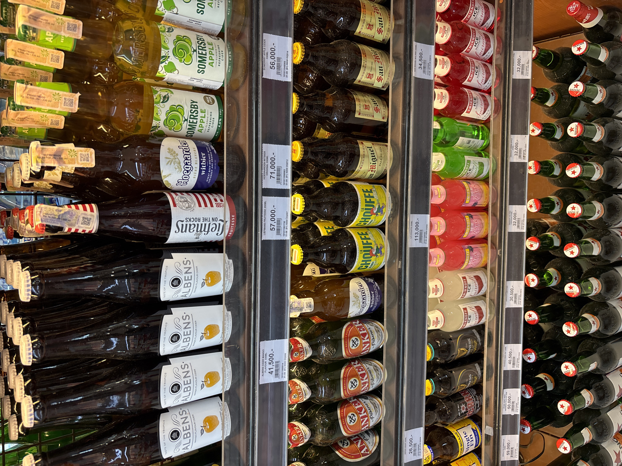 This image captures a well-stocked beverage aisle within a store, likely a supermarket or liquor store, in Kuta Selatan, Indonesia. The shelves are filled with numerous bottled drinks, predominantly alcoholic beverages, neatly arranged for sale.
Visible products include multiple rows of Somersby Apple Cider, Hoegaarden Witbier, Liefmans Fruitesse, and Albens Mango Flavour Cider on the left. The middle section features San Miguel Pale Pilsen, La Chouffe, and Anker Pilsener. On the right, shelves hold Smirnoff Ice (in various flavors like pink and clear), Guinness Foreign Extra Stout, Single Fin Summer Ale, and a prominent display of Bintang beer bottles.
Each product has a visible price tag, indicating prices in Indonesian Rupiah (e.g., 56,000.- for Somersby Apple Cider, 71,000.- for Hoegaarden Witbier, 41,500.- for Albens Cider, and 32,000.- for Bintang). Some tags also show product codes, volumes (e.g., 330ML, 250ML), and expiry/batch information. "New Look" is noted on some Somersby bottles, indicating a packaging update. Alcohol content (ALC. 4.5% VOL.) is also visible on some labels. The general atmosphere suggests a commercial setting during operational hours. No people are visible, focusing entirely on the products and their display.