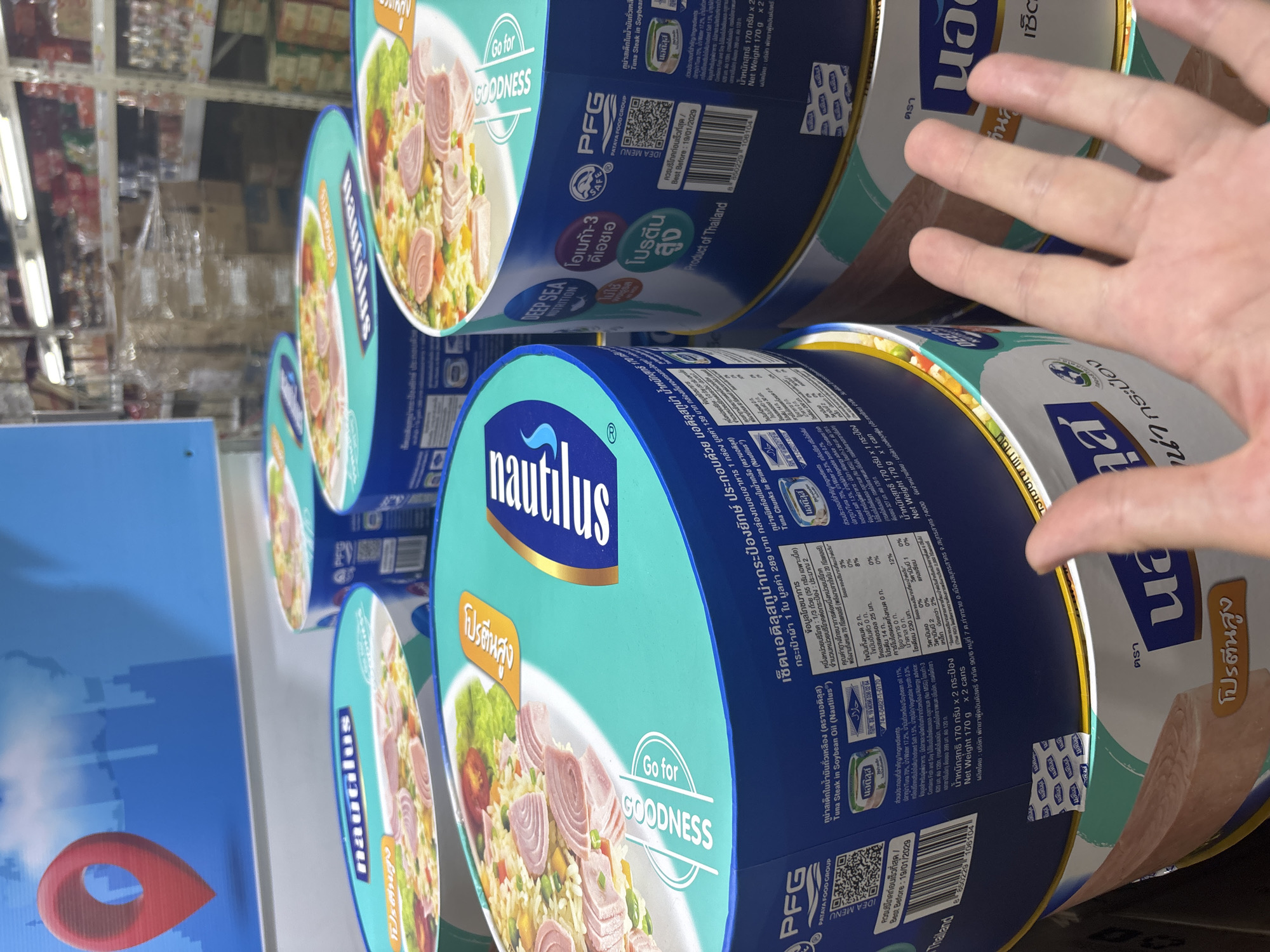 The photograph presents a view of several cans of "Nautilus Tuna Steak in Soybean Oil", a product of the Pataya Food Group (PFG), stacked on a shelf. The cans feature a light blue background with images of a salad-like dish and text in Thai and English. The labels include nutritional information, ingredients, and a "Best Before" date of 19/01/2029. A hand is visible in the foreground. The cans are likely located in a store in the city of Baan Bo Phut, Thailand.