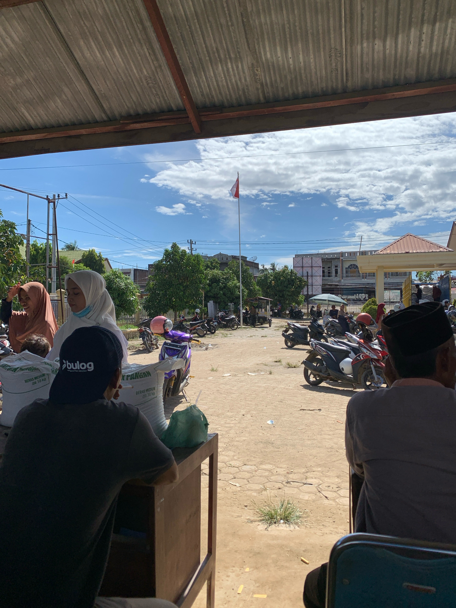 The image depicts an outdoor scene, likely a public area, in Pantegurah, Indonesia, under a partly cloudy sky. A corrugated metal roof is visible in the foreground, providing shade. In the mid-ground, several people are gathered. Some individuals are wearing headscarves. There are stacks of large bags, possibly containing rice, with text including "BERAS MEDIUM" and "10 Kg". The logo "bulog" is visible on one of the bags. Various motorcycles are parked around the area, including a purple scooter and several others. A flag on a pole, appearing to be the Indonesian flag, is visible in the distance. Buildings and trees form the backdrop of the scene. Two men are seated in the foreground, facing away from the camera. One man is wearing a dark cap with a logo and appears to be smoking. The ground is sandy and uneven, with some patches of vegetation and scattered debris.
