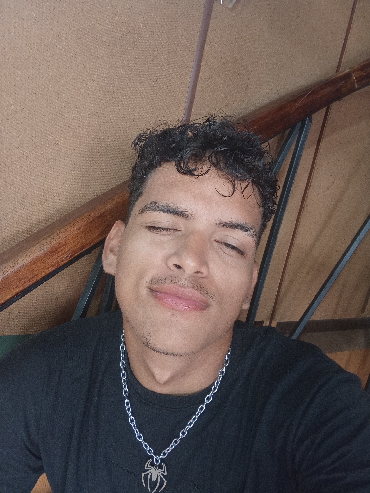 A person is posing for a selfie indoors, with a brown paneled wall behind them and the side of a wooden staircase railing visible in the background. The person has dark, curly hair, lightly tanned skin, and is wearing a black t-shirt and a silver chain necklace with a spider-shaped pendant. The person is looking directly at the camera with one eye closed and a slight smile. There is no information to confirm the location is in Juan Griego, Venezuela.
