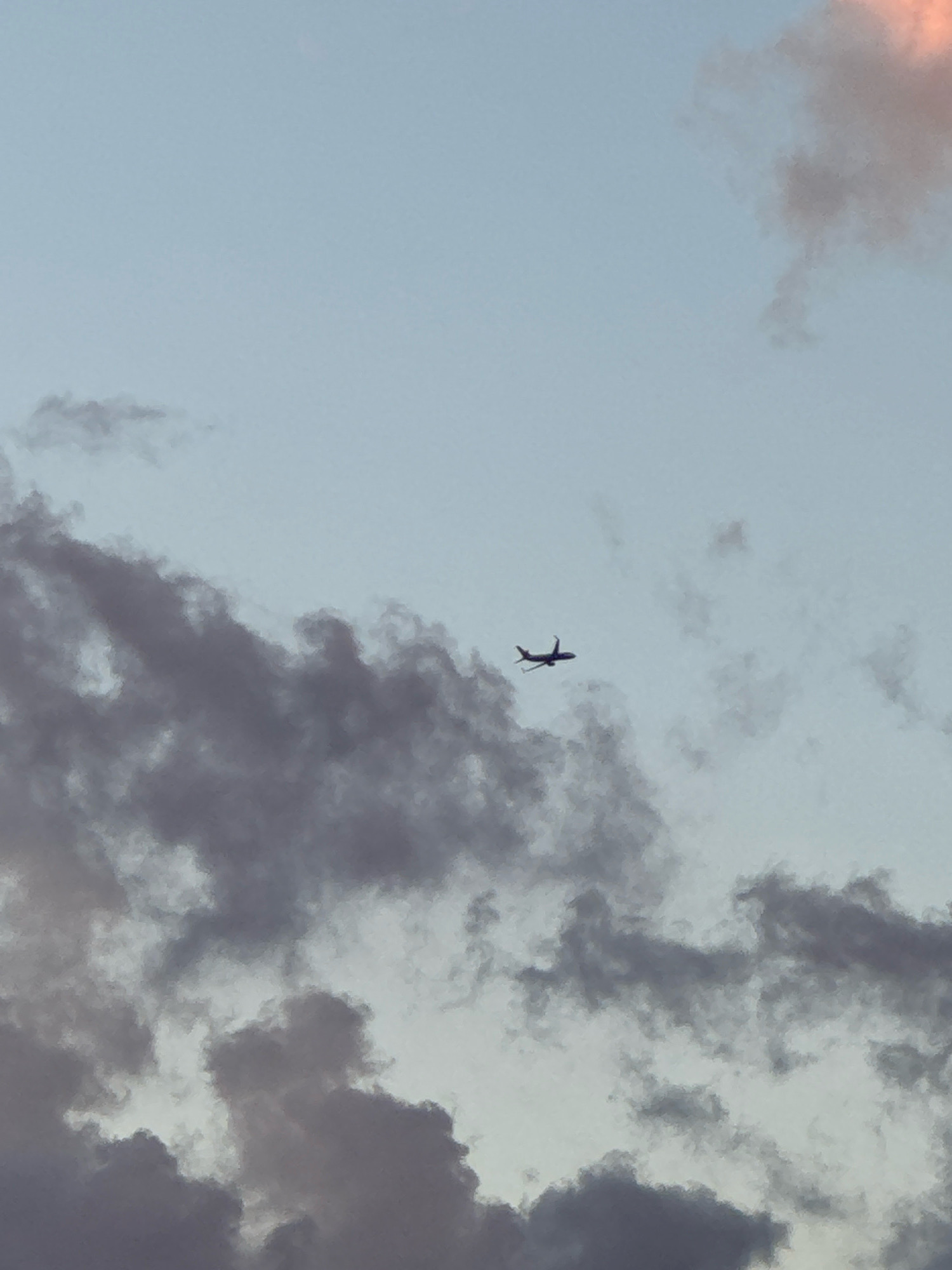 An airplane is silhouetted against a pale blue sky with scattered clouds in Capaci, Italy. The aircraft, seen from the side, is in mid-flight, with its wings extended. Dark gray clouds occupy the lower portion of the frame, with some lighter clouds higher up and a hint of pinkish-orange cloud in the upper right corner, suggesting the time may be near sunset.