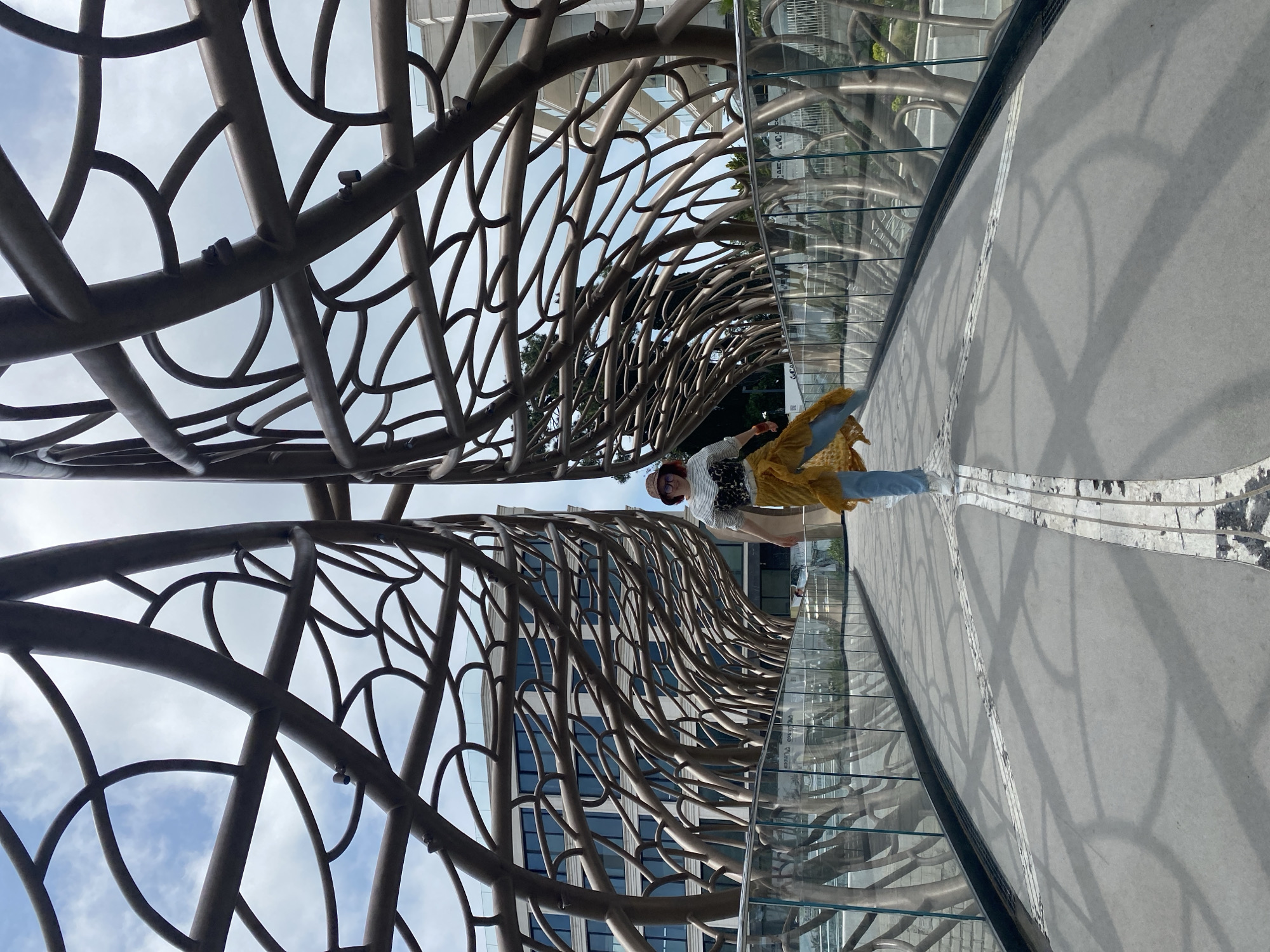 The image shows a woman standing on a pedestrian bridge in Sochi, Russia. The bridge features a unique, ornate metal framework overhead, resembling interwoven branches. The bridge's sides are lined with glass panels. The pedestrian surface is light gray concrete. A building with blue-tinted windows is visible in the background. The sky is partly cloudy. The woman is wearing a hat, a striped shirt, light blue pants, and a flowing, gold-colored jacket or wrap.
