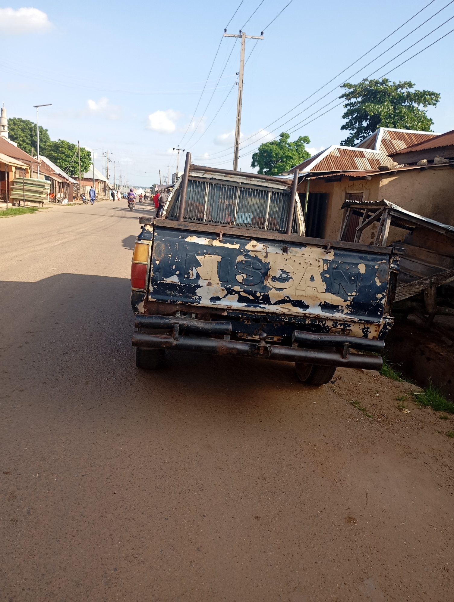 In Shendam, Nigeria, a street scene unfolds under a partly cloudy sky. The foreground is dominated by the rear of a dark blue pickup truck with significant wear and peeling paint. The truck's tailgate displays faded yellow lettering, partially obscured, but appearing to spell out "NISSAN." A metal bumper with dual exhaust pipes is visible. To the right, a grassy embankment slopes upward, adjacent to a rough, wooden structure with a corrugated metal roof. The street itself is paved and appears to be a thoroughfare in a town or village. Along the left side of the street, a row of buildings with corrugated metal roofs line the road, some appearing to be shops or residences. A minaret of a mosque is visible in the distance. Several people are present, with some individuals riding motorcycles and others walking along the street. The environment suggests a typical day in a Nigerian community.