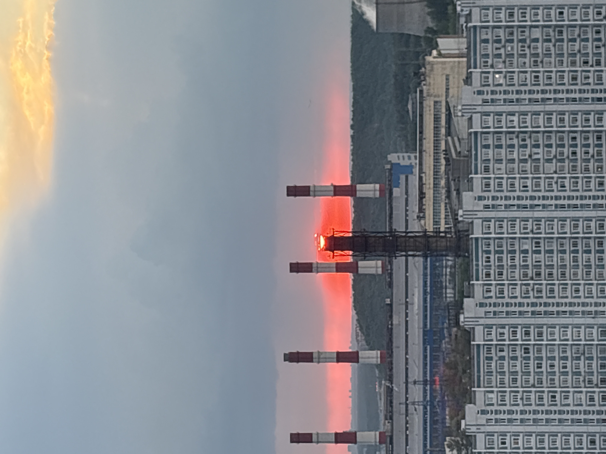 The image shows a cityscape at dusk or dawn. The sky is divided between a cloudy, pale blue and an area of orange light, possibly from the sun. Several industrial smokestacks with red and white bands stand prominently in the middle ground, some with a visible red glow at their tops. Behind the stacks, there are trees. To the right of the image, there is a tall building with many square windows, suggesting an apartment or office structure. There are multiple train tracks next to the building.