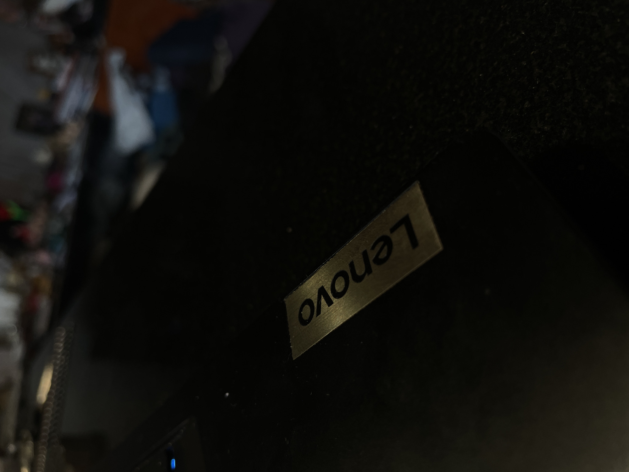 The image shows a close-up of a black electronic device, likely a laptop or monitor, with a silver "Lenovo" logo affixed to its surface. The logo is oriented diagonally in the frame. The background is blurred and shows a cluttered area with various objects. The lighting is dim, highlighting the Lenovo logo. There are no people visible in the frame. There is no evidence to suggest this image was taken in Burgas, Bulgaria.