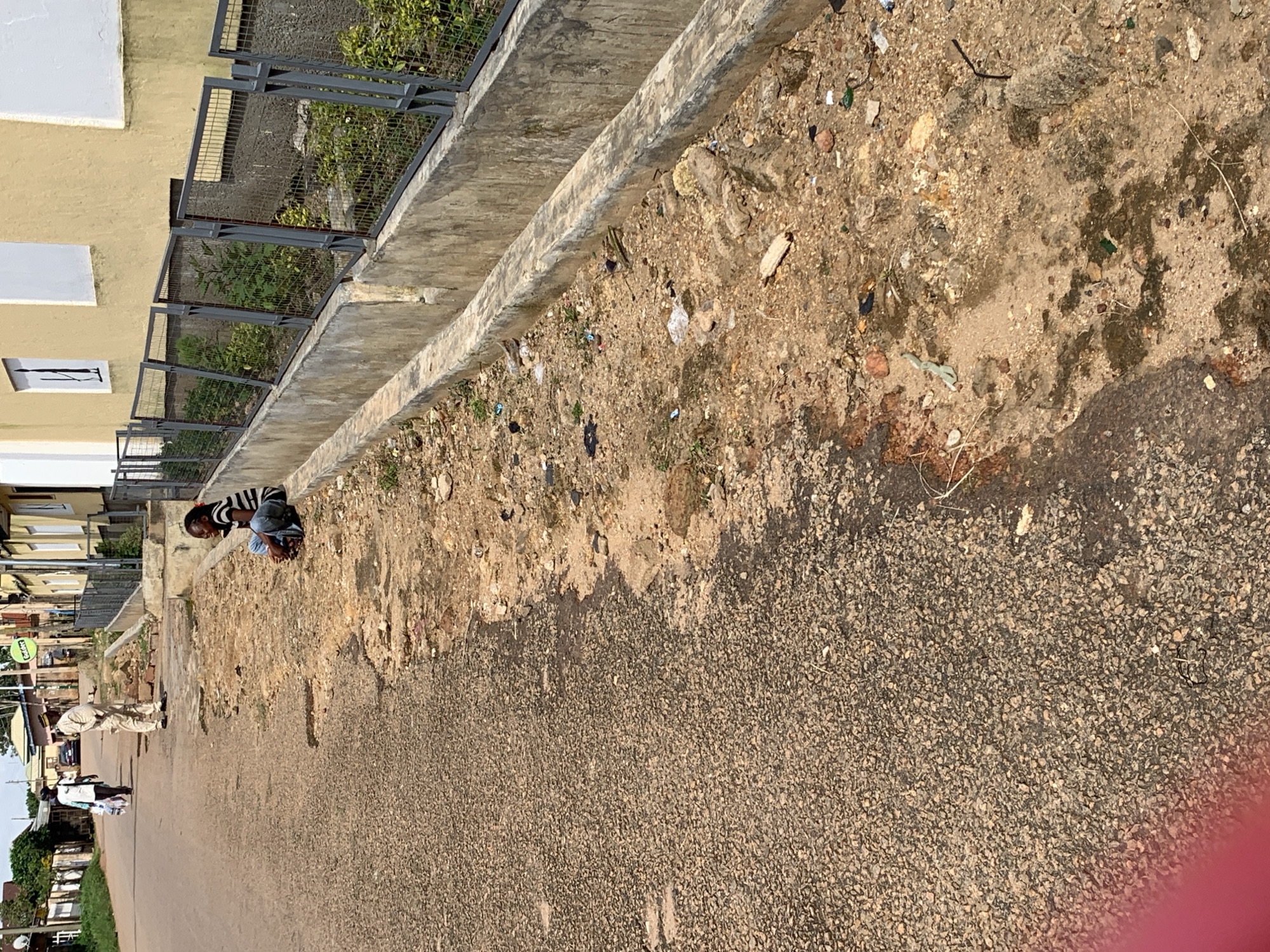 The image depicts a street scene in an urban environment. A young person wearing a striped shirt and blue jeans is sitting on a concrete curb next to a drainage ditch filled with debris. The ditch is adjacent to a paved road. In the background, two individuals are walking away from the camera, one carrying shopping bags. Further down the street, buildings with yellow and white facades are visible, along with a metal fence. The overall setting appears to be a moderately developed urban area with signs of neglect, indicated by the trash in the ditch and the worn road surface. The lighting suggests it is daytime, possibly mid-morning or afternoon.