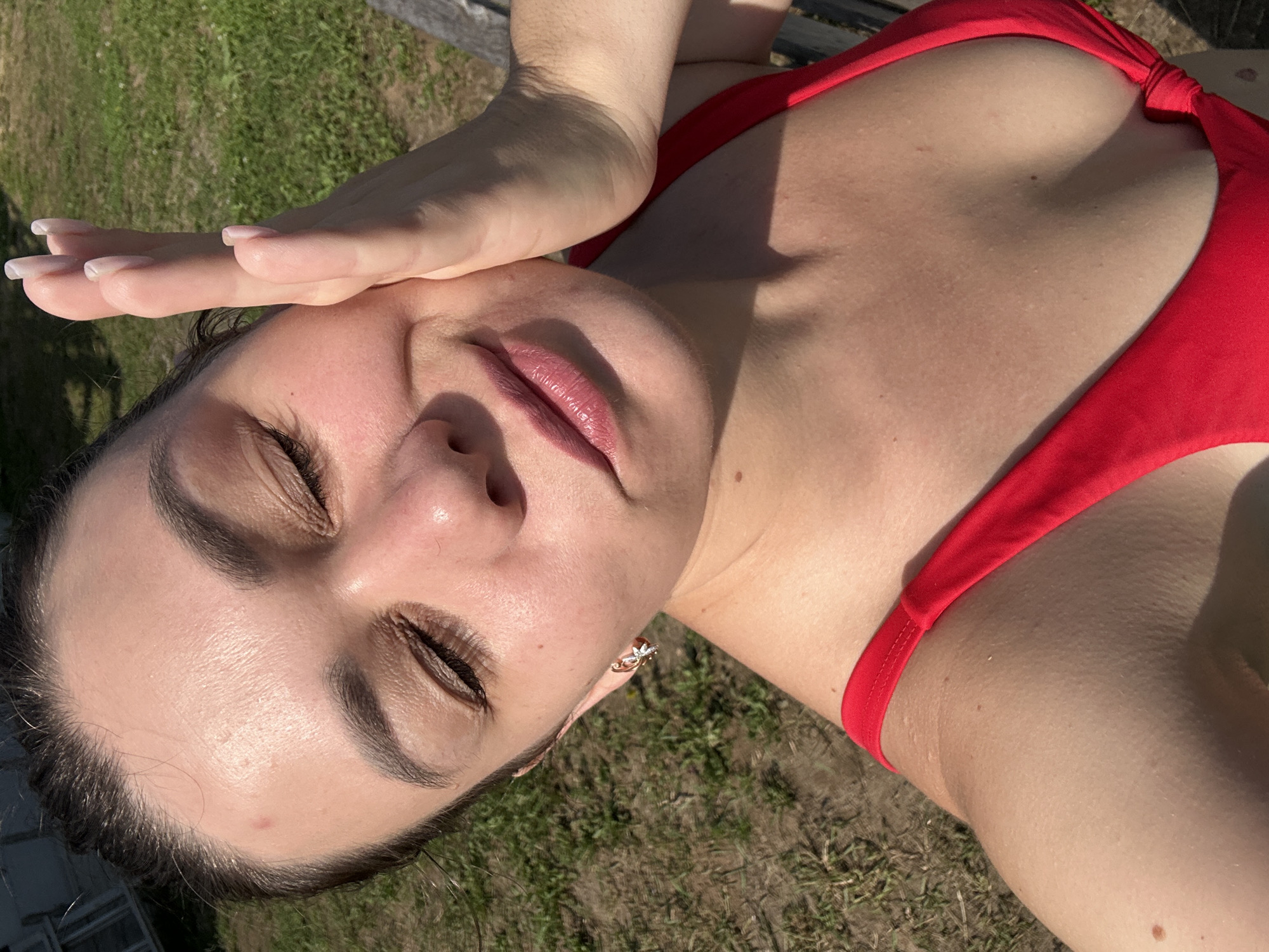 A woman in a red swimsuit is taking a selfie outdoors on a sunny day. She has her eyes closed and is touching her cheek with her right hand. The background shows green grass and some wooden structures. The image appears to be taken at ground level. There is no indication of the specific location within the city of Chaikovsky, Russia.
