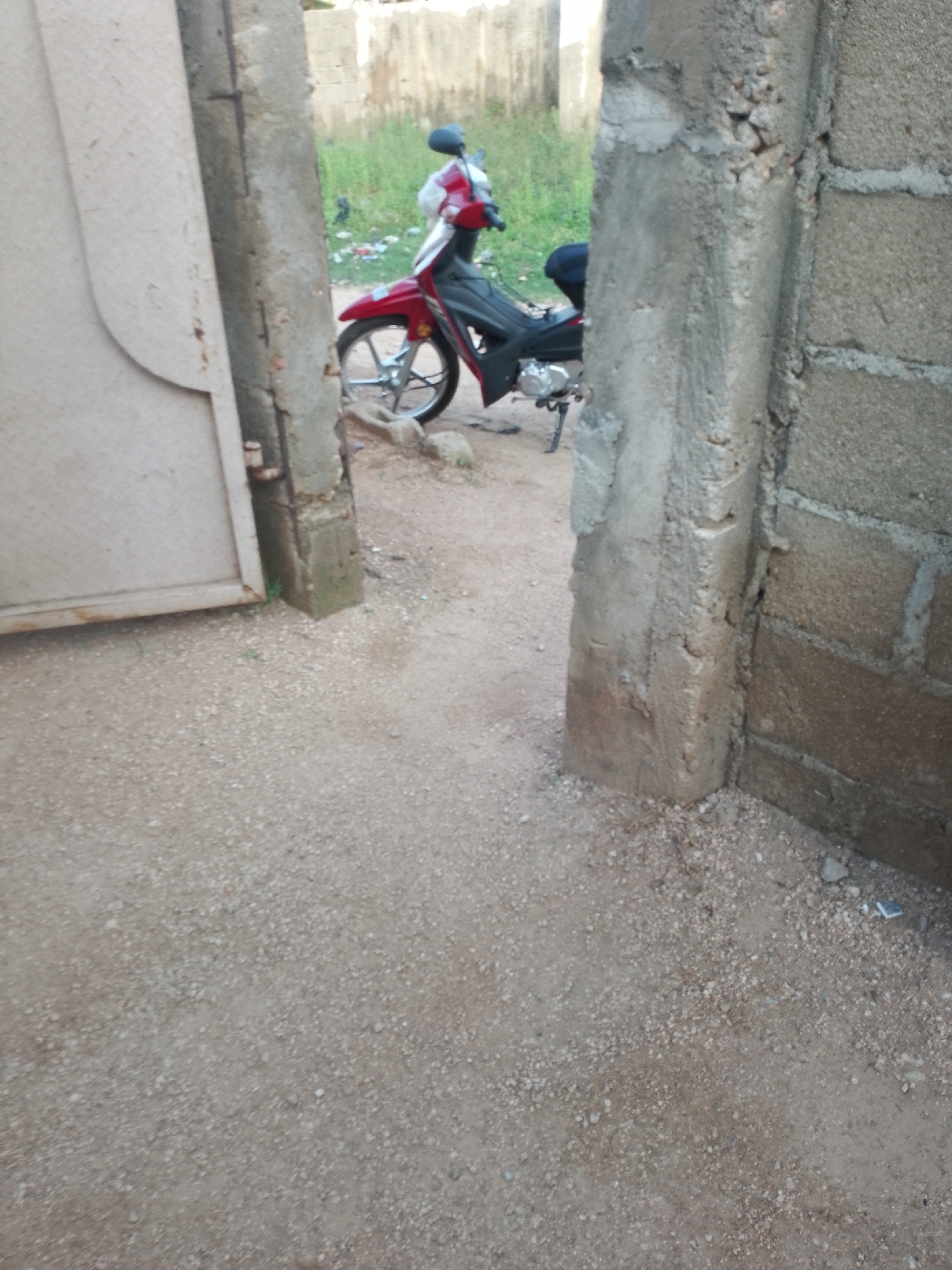 A red and black motorcycle is parked in a dirt yard, partially obscured by a concrete pillar and a beige metal gate. The motorcycle has a black seat and fenders, and a silver engine. It is facing to the left of the frame. Behind the motorcycle, there is a patch of green grass and some debris. The scene appears to be in an outdoor, possibly urban, environment with rough ground composed of dirt and small stones. The gate is to the left of the frame, and a concrete wall is on the right.