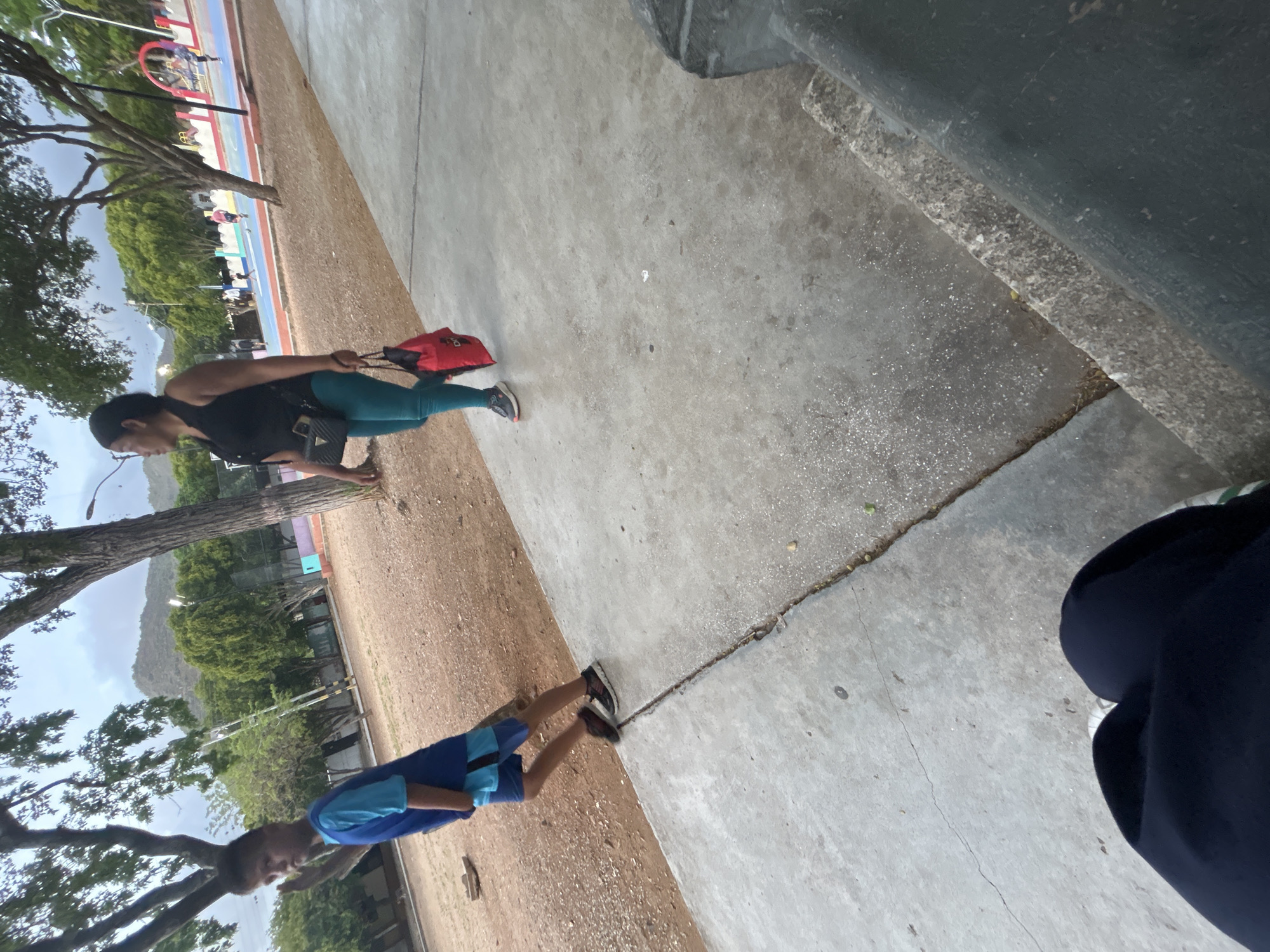 In Santa Ana, Venezuela, a boy in a blue shirt and shorts walks along a concrete path. A woman in a black sleeveless top and teal leggings walks behind him, carrying a red bag in her right hand and a black purse on her left shoulder. The ground is a mix of dirt and concrete, with trees and a playground visible in the background. The sky is overcast, and mountains can be seen in the distance.
