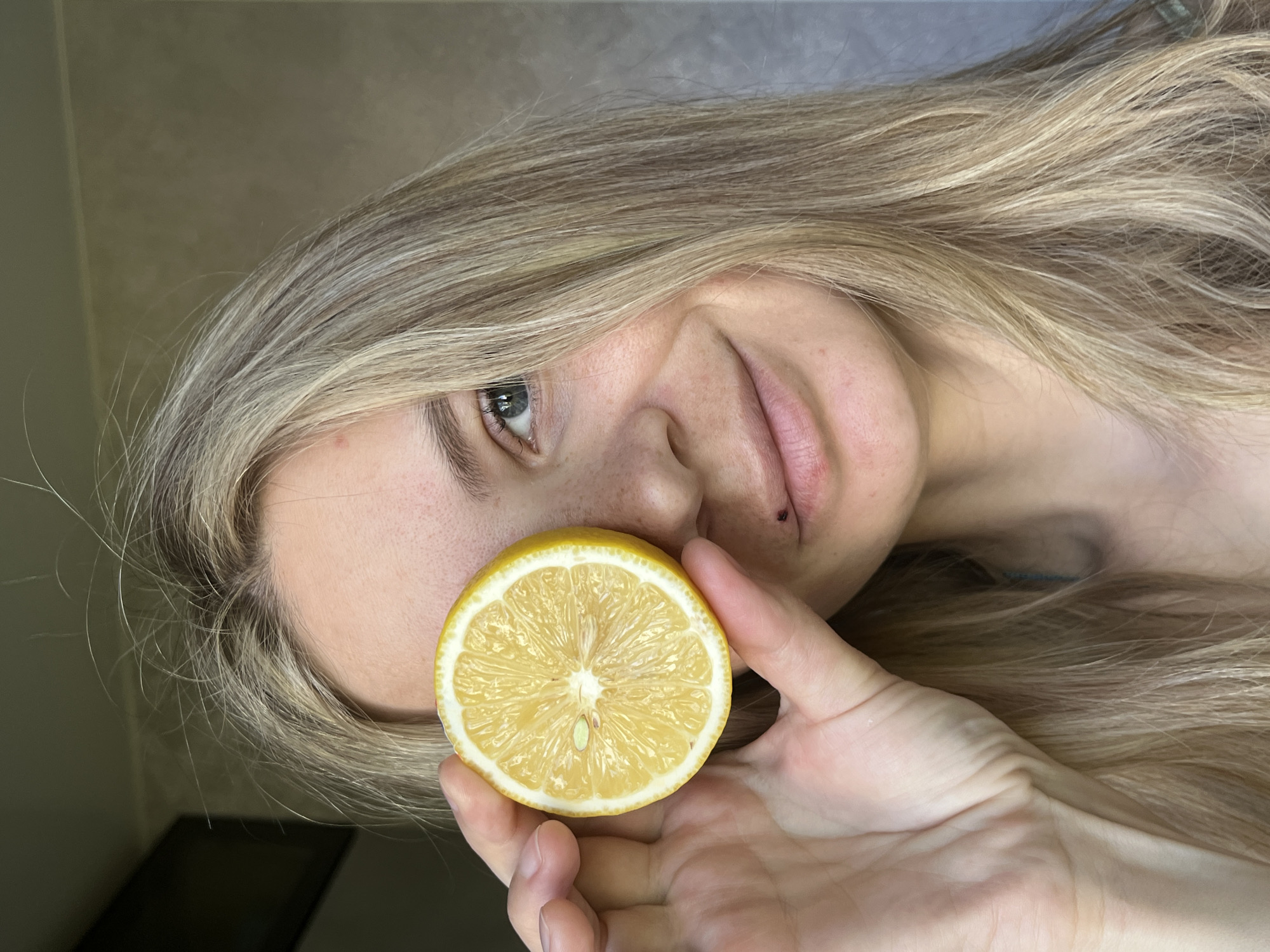 A woman with long blonde hair holds half a lemon in front of her face. Her left eye is visible. The background is blurry and indistinct. The photograph appears to have been taken indoors. There is no indication in the image that would reveal its location, including whether it was taken in Chaikovsky, Russia.