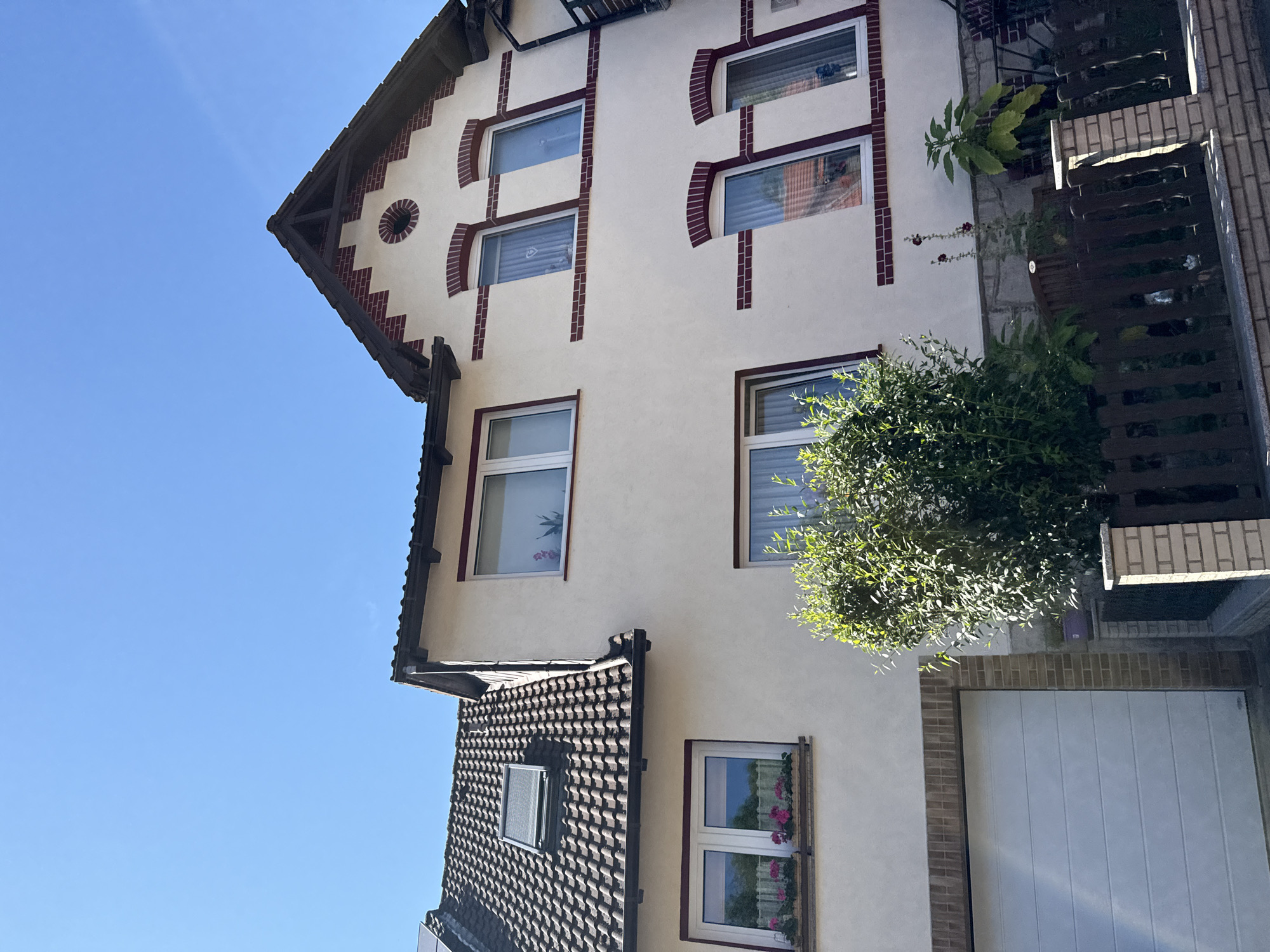 The image shows a multi-story residential building with a tiled roof, situated in the city of Bad Sachsa, Germany. The building features white stucco walls with decorative dark red brick accents around the windows and along the roofline. Several windows are visible, some with flower boxes containing pink flowers. A white garage door is present on the ground floor. A lush green bush partially obscures the lower part of the building, and a wooden fence runs along the front. The sky above is clear and blue.