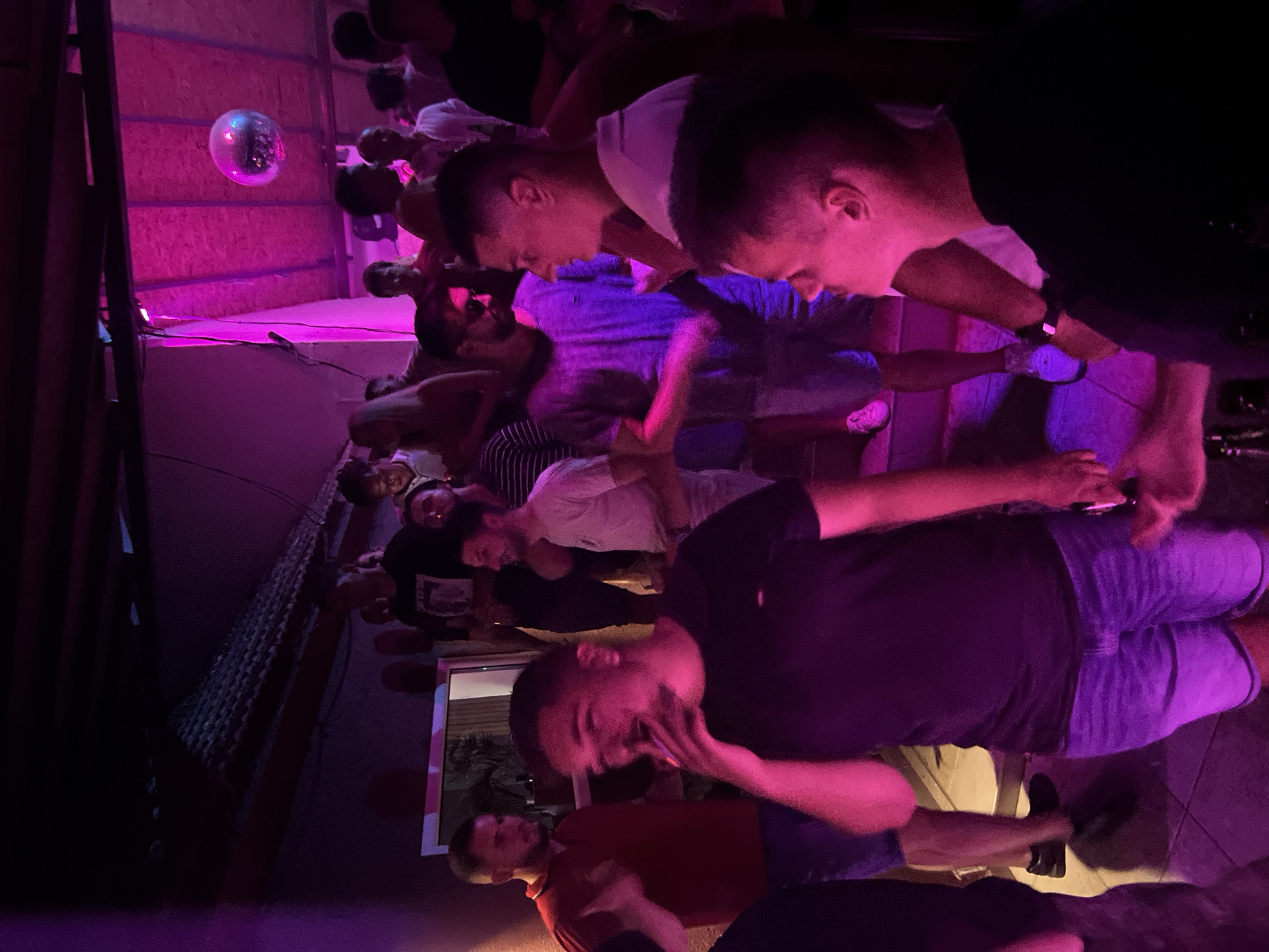 The image depicts a dimly lit nightclub or bar scene, characterized by vibrant pink and purple lighting. In the foreground, a man in a black t-shirt and denim shorts is the central figure, with his face partially obscured as he appears to be smoking. To his left, a man in a red polo shirt and dark pants stands with his arm raised. Behind them, several other individuals are visible, some engaged in conversation or dancing. A disco ball hangs from the ceiling, reflecting the ambient light. The overall atmosphere suggests a lively social gathering. The location is identified as Herceg Novi, Montenegro.