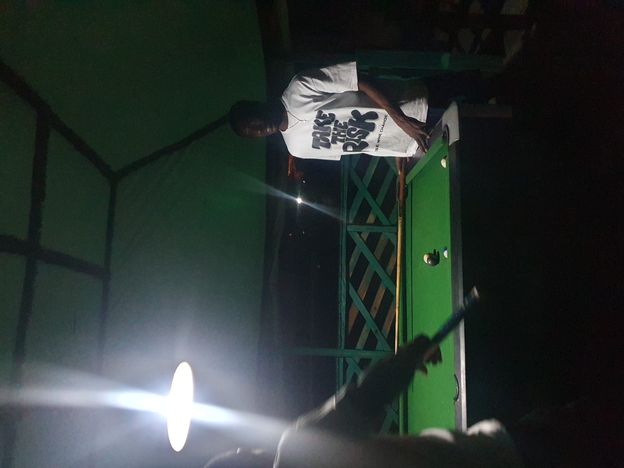 This image shows two individuals at a dimly lit outdoor pool table, possibly a bar or recreational area, in Lokoja, Nigeria. A bright overhead light illuminates the scene. One person, wearing a white t-shirt with the text "TAKE THE RISK" and a cross pendant, stands by the pool table, their hands near the edge. Their facial expression is obscured by the shadows and light. The other person, partially visible in the foreground, is holding a pool cue, appearing to be in the process of playing. The pool table is covered in green felt, with several balls on it, including a striped ball and a white cue ball. The background reveals a green wooden lattice structure and dark foliage, suggesting an outdoor setting. The overall atmosphere is casual and focused on the game.