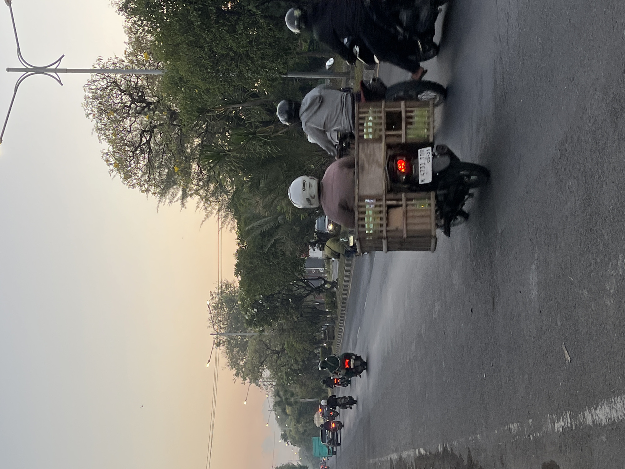This is a photograph taken on a street in Ngaban, Indonesia, likely during early morning or late afternoon given the soft, warm light. The scene depicts a road with several motorcyclists. In the foreground, a motorcycle with a wooden cargo carrier is visible, carrying a passenger with a white helmet. Behind this, other motorcyclists are also present, some with passengers. Trees line the side of the road, and streetlights are visible in the distance. The sky is a pale orange and yellow hue. The road appears to be asphalt and is wet in some areas, suggesting recent rain. The overall atmosphere is calm and captures a typical daily scene of transportation in the city.