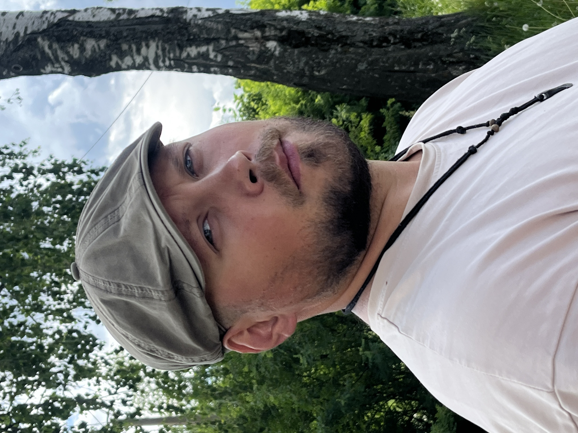 A selfie of a man wearing a newsboy cap and a light-colored shirt with a beaded necklace or lanyard. He has blue eyes, a beard, and stubble. The background includes a tree branch and foliage, suggesting an outdoor setting. The image is rotated, making it appear as if the man is looking upward. It is not possible to verify the location.