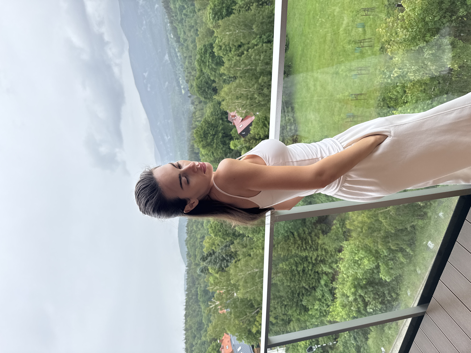A young woman is captured in a vertical, full-body shot, standing on a balcony or terrace and gazing out at a scenic landscape. She has dark, medium-length hair, styled away from her face, and is wearing a light-colored, possibly white or off-white, sleeveless top and matching wide-leg pants or a jumpsuit. Her left hand is casually placed in a pocket of her pants. Her head is tilted slightly upward, and her eyes appear closed or downcast, giving her a serene and contemplative expression.
The balcony features a modern design with a glass railing and dark, slim metal supports. The floor of the balcony is made of light-colored, possibly composite, decking material.
Beyond the balcony, a vast, verdant landscape unfolds. Rolling hills densely covered with lush green trees dominate the midground. In the distance, towering mountains rise under a soft, overcast sky, which appears cloudy and diffused. Hints of red-roofed buildings can be spotted nestled among the trees in the valley below, suggesting a rural or semi-rural setting. The light suggests an overcast daytime, and there might be moisture on the glass railing, hinting at recent rain or high humidity.
No visible text is present in the image.