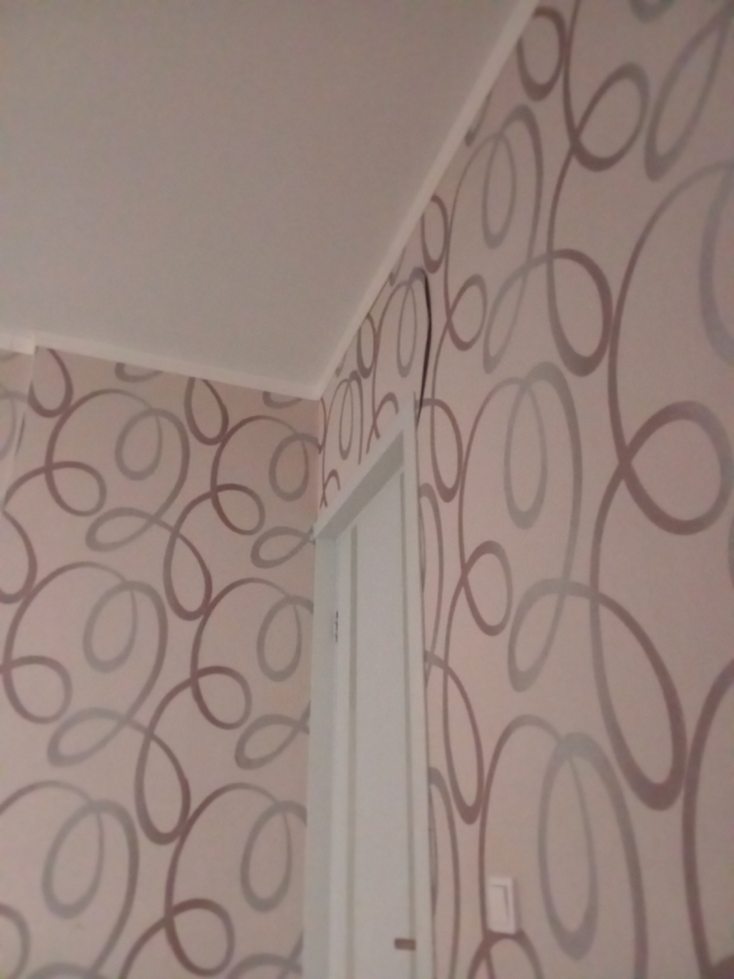 This is an eye-level, medium shot of a room interior, focusing on a corner where two walls meet. The walls are covered in wallpaper with a repeating pattern of light brown, swirling lines. A white ceiling is visible in the upper left quadrant of the image. On the right side, a portion of a white doorframe is visible, with a white door jamb. A light switch is mounted on the wallpapered wall to the right of the doorframe. The scene appears to be within a residential dwelling. The location in the city of Kazan, Russia cannot be determined from the image.
