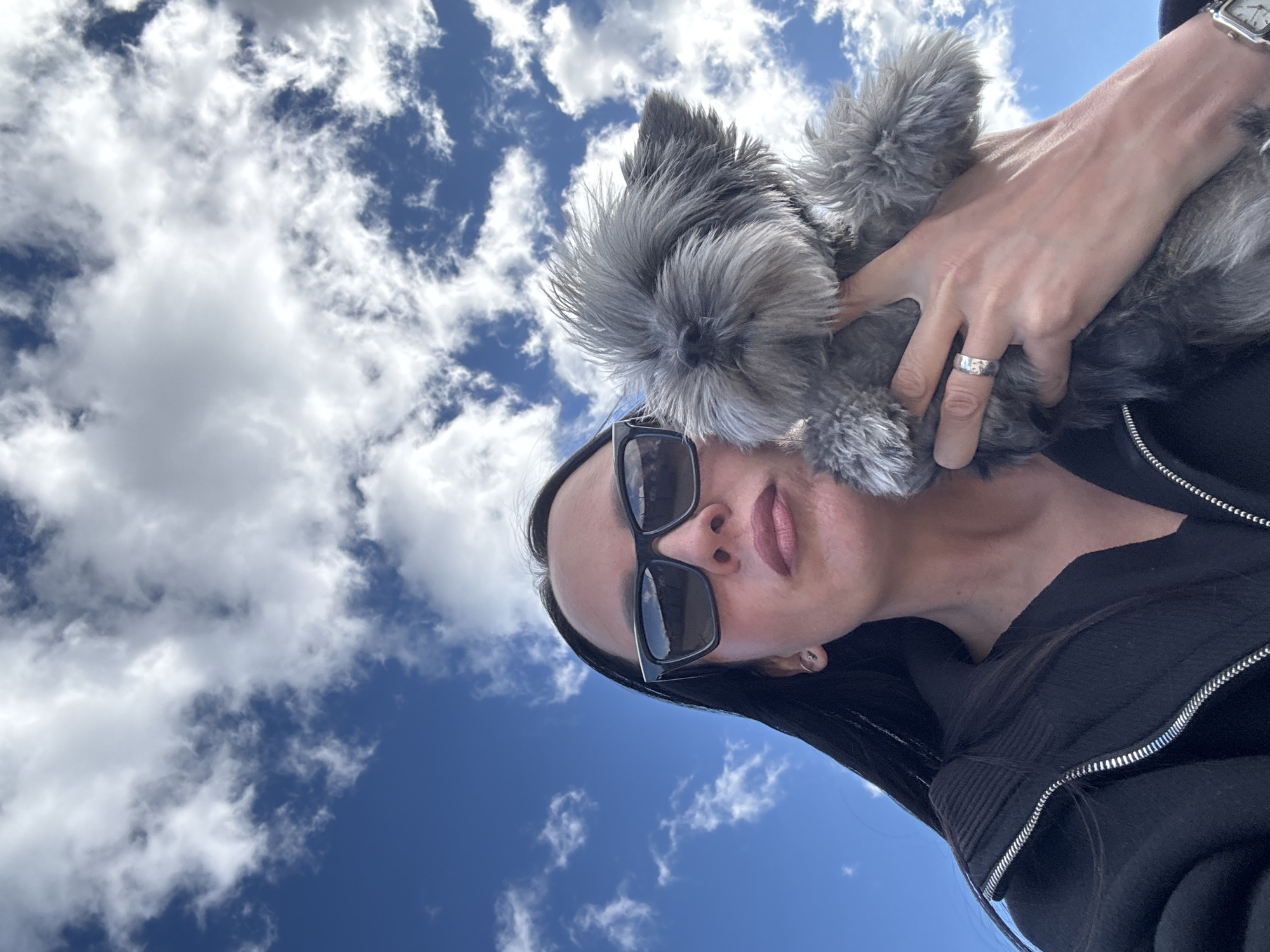 A low-angle selfie shows a person holding a gray, small dog. The person is wearing sunglasses and a dark-colored zip-up jacket, and is wearing a ring on their left ring finger and a watch on their left wrist. The sky is blue with scattered white clouds. There are no identifying indicators of the city of Chaikovsky, Russia.