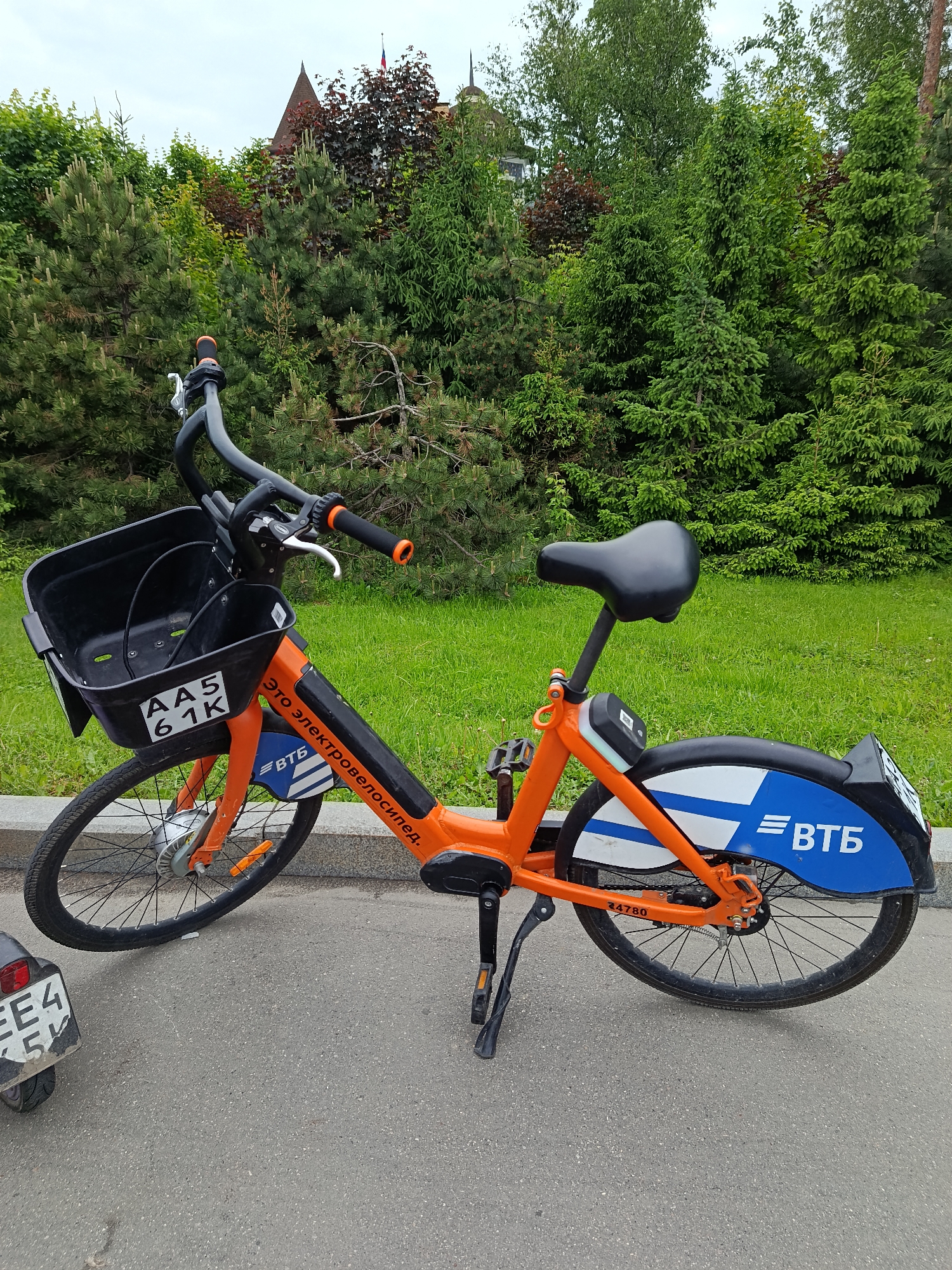 An orange and blue electric bicycle is parked on an asphalt path next to a grassy area, surrounded by numerous green trees under an overcast sky. The bicycle, a model from a shared rental system, is oriented with its front wheel to the left.
The bike's frame is a bright orange, complemented by black handlebars, a black seat, and a large black plastic basket mounted on the front. On the front basket, a white plate with black text reads "AA5 61K". The main frame displays white Russian text: "Это электровелосипед." (meaning "This is an electric bicycle."). The rear mudguard is blue and white, prominently featuring the "ВТБ" (VTB) logo, indicating sponsorship. A white number "24780" is visible on the rear of the frame near the wheel. The front wheel includes a silver hub, likely housing the electric motor. A black kickstand is deployed, holding the bike upright.
The background is rich with greenery, featuring various species of trees including tall pines, spruces, and other deciduous trees, suggesting a park or well-maintained landscape. Peeking through the trees in the upper-center is the dark, angular roof of a building with a spire, and a small flag with white, blue, and red horizontal stripes (the Russian flag) is visible at its apex. A small portion of another vehicle, possibly a scooter or moped, with a partial license plate "ЕЕ4 5" is visible in the bottom left corner.
The image captures an outdoor scene, likely in Moscow, Russia, during daytime under natural, diffuse light. The bicycle is stationary, and no people are present.
