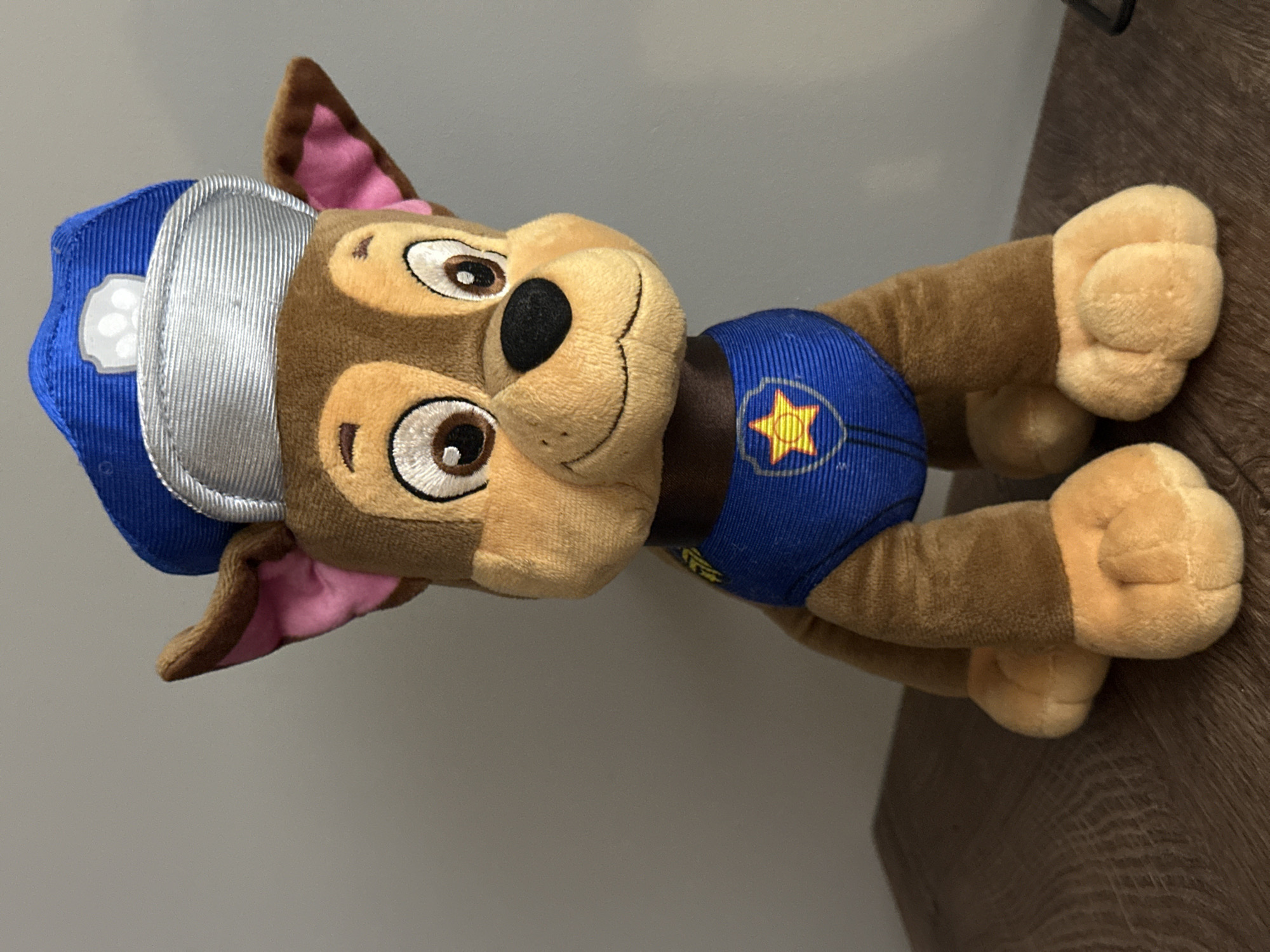 The media file displays a plush toy of Chase from the animated series Paw Patrol, presented both as a full, rotated image and several close-up crops. The toy is primarily brown and tan, featuring a blue police cap with a silver, ribbed visor and a white paw print emblem on the side. Its inner ears are a contrasting pink. The toy's face shows large, embroidered eyes with white pupils, a black nose, and a stitched smiling mouth. It wears a blue, ribbed uniform around its torso, detailed with a yellow star badge with a red outline.
In the full image, the toy is lying on its side, positioned against a plain, light grey wall in the background and resting partially on a brown, wooden surface, which appears to be a table or shelf with visible wood grain. The close-up crops highlight various features: one focuses on the blue hat, silver visor, white paw print, and part of the face; another shows the toy's smiling face, black nose, and the yellow star badge on its uniform; a third crop details one of the plush, tan paws resting on the wooden surface; and other crops provide views of the pink inner ear and portions of the body and background.
There are no people visible in the images, and no specific activity or event is taking place beyond the static display of the toy. The setting is indoors, with ambient lighting, and no discernible time of day or specific weather conditions can be determined. The visible text is limited to the design elements on the toy's uniform and hat (paw print, star). The overall scene is a simple display of a children's character toy, located in Bobov Dol, Bulgaria.