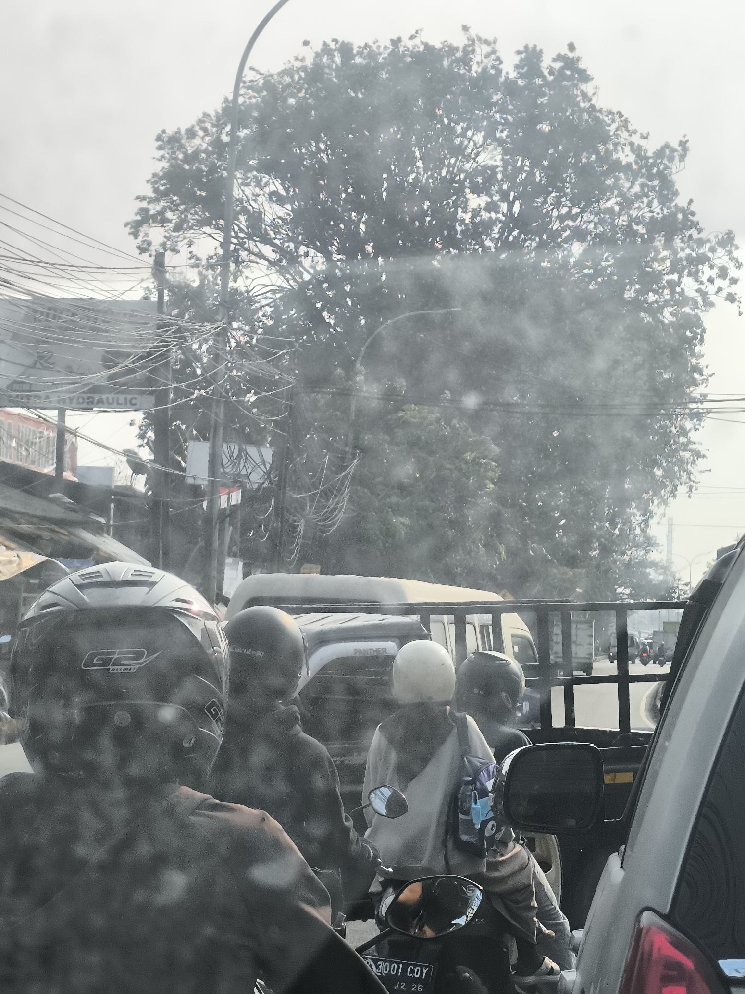 The image depicts a street scene in Tangerang, Indonesia, likely during daylight hours given the illumination. In the foreground, from a rear perspective, are two individuals on a motorcycle. The person in the front is wearing a black helmet with a "G2 HELMET" logo and a patterned shirt. Behind them, another person is visible, also wearing a dark helmet, their face obscured. They appear to be traveling behind other vehicles.
Further into the scene, several other individuals are present, primarily on motorcycles, suggesting a busy urban environment. One motorcycle in the mid-ground carries at least two passengers wearing helmets, one white and one black. They are positioned in front of a utility vehicle or truck bed, which is fenced. Another truck with a similar fencing structure is visible behind them, and further back, several more motorcycles are seen navigating the road.
The surrounding environment includes dense overhead power lines, characteristic of urban infrastructure, and a large tree with extensive foliage on the right side of the frame. Buildings and storefronts are partially visible on the left, with signage that appears to read "HYDRAULIC". The overall atmosphere suggests a typical traffic condition in a bustling Indonesian city. The image is slightly hazy, possibly due to atmospheric conditions or a dirty lens.
