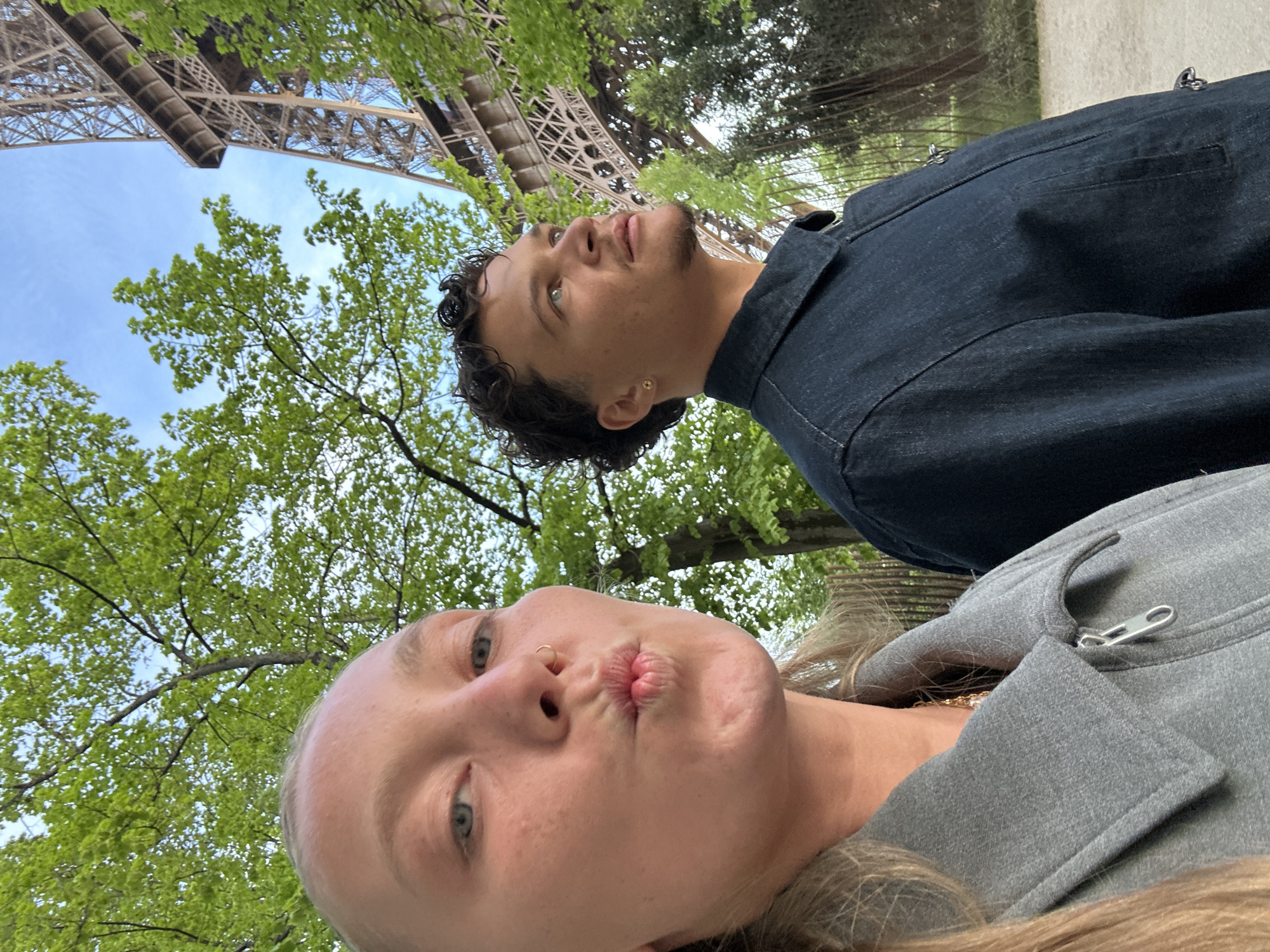 The photo depicts two individuals in Paris, France, with the Eiffel Tower visible in the background. The foreground features a blonde woman puckering her lips for the camera. Behind her, a man with curly hair gazes upward. The environment includes trees with green foliage, suggesting a park or garden setting near the iconic landmark.