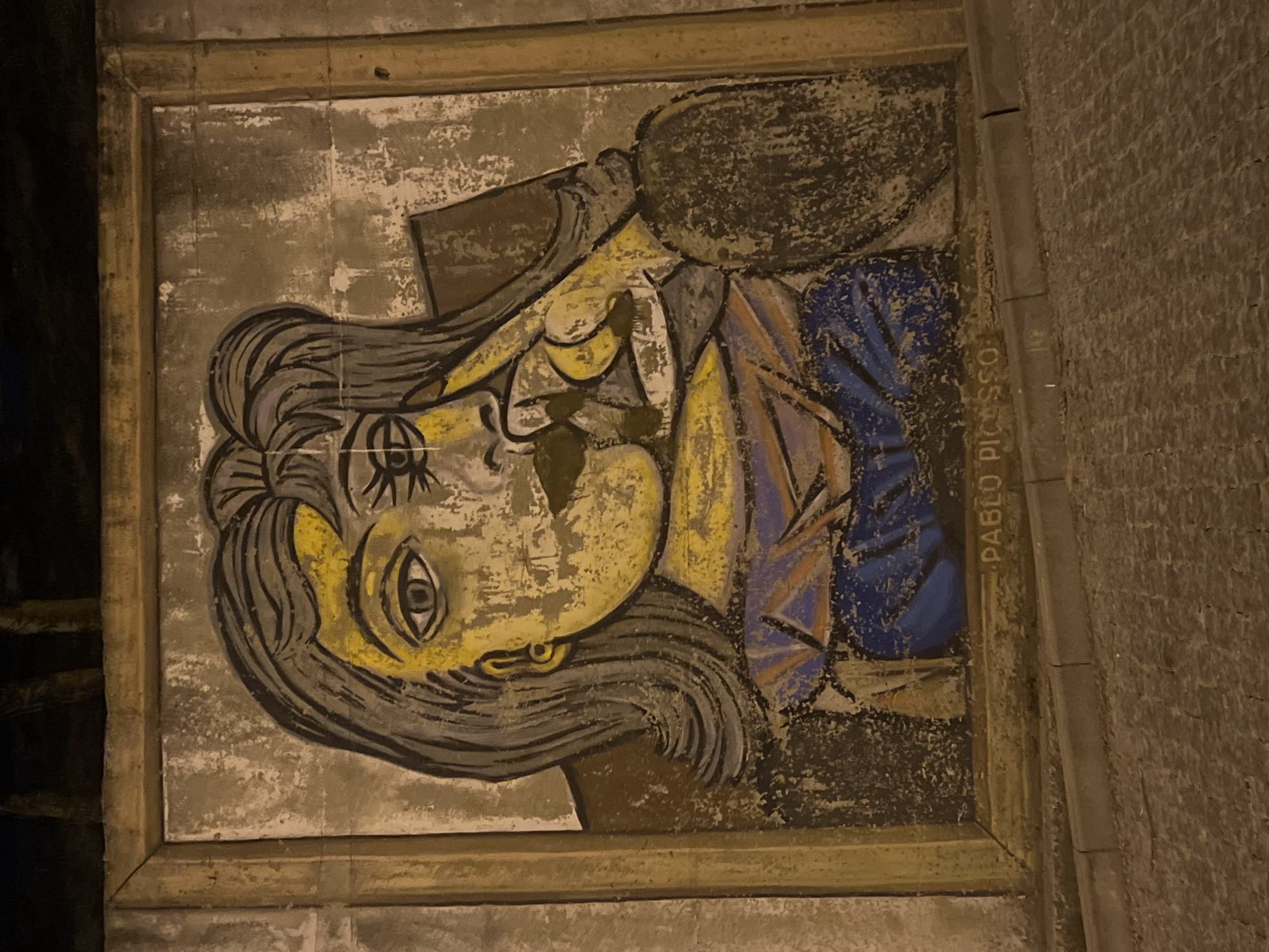 The image displays a mural of a woman's face, rendered in a cubist style reminiscent of Pablo Picasso's work, prominently displayed on a textured wall. The mural features bold outlines and fragmented features, with a yellow hue dominating the face, accented by blue and purple clothing. The word "PABLO PICASSO" is inscribed in capital letters along the bottom edge of the mural. The mural is framed by a weathered, light brown border. The wall itself appears aged and chipped in places. The foreground shows cobblestone pavement and a concrete curb, indicating an outdoor urban setting. In the background, dark, out-of-focus shapes suggest trees or other structures against a dark sky. The lighting appears to be artificial, casting a warm glow on the mural. This mural is located on the wall of a building in the city of Odesa, Ukraine.