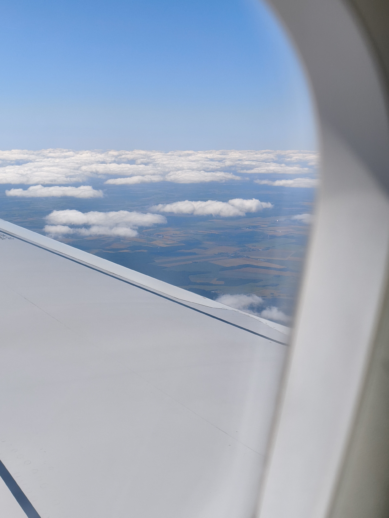 The image is an aerial view from an airplane window, looking down at a landscape. The sky is a clear, bright blue. Below the sky, a layer of white, fluffy clouds stretches horizontally across the frame. Beneath the clouds, the ground is visible, with a patchwork of agricultural fields in various shades of green and brown. There are some trees and buildings scattered across the landscape, suggesting a rural or semi-rural area. An airplane wing, a prominent white structure, occupies the lower left portion of the image. The right side of the frame is taken up by the interior of the airplane window frame, which is a neutral gray color. There are no people or specific landmarks visible that would identify a precise location.