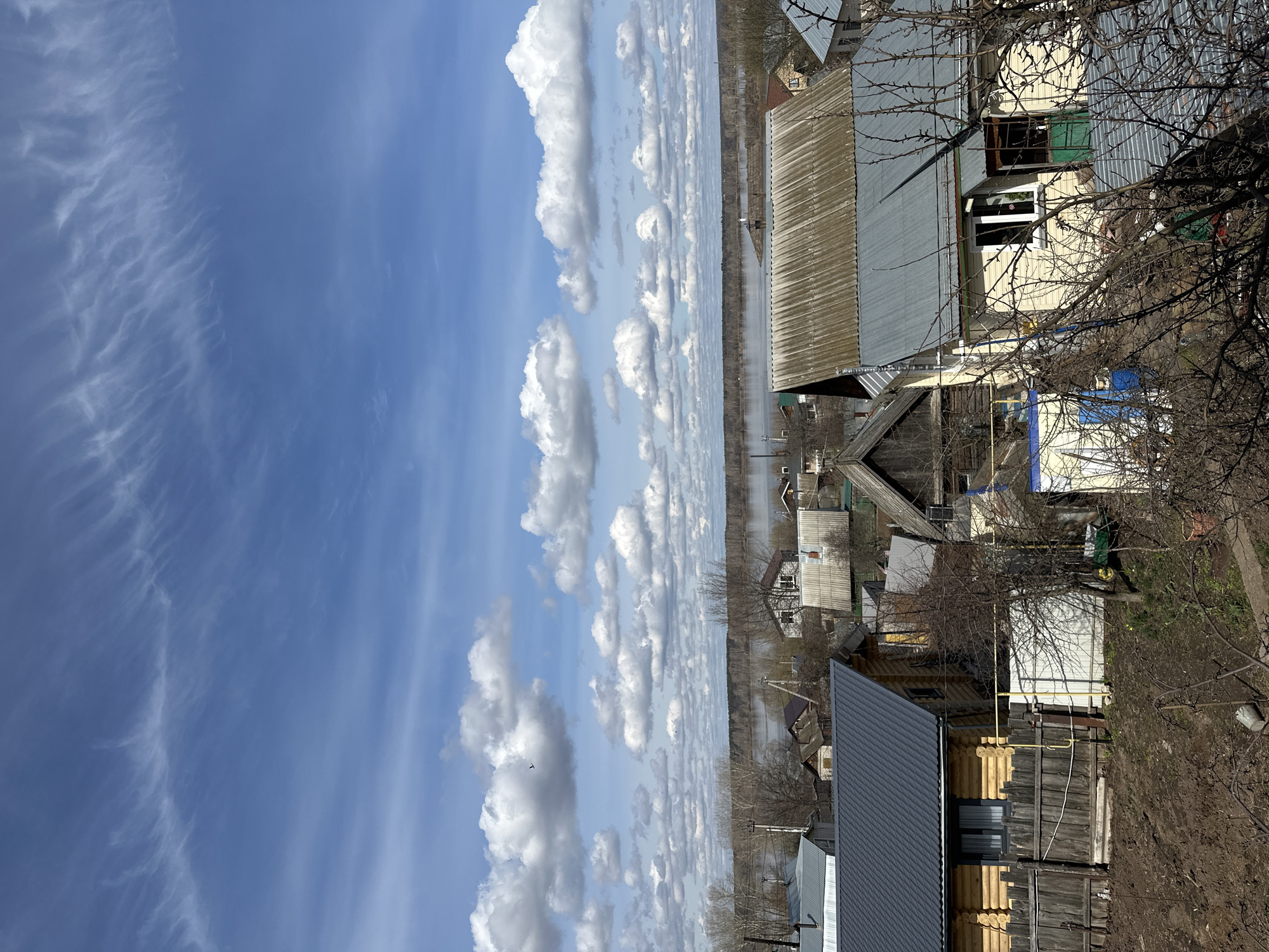 The image displays a panoramic view of a rural or semi-urban landscape under a bright sky, likely during late winter or early spring. The perspective is elevated, looking down towards a settlement and a body of water.
The upper portion of the scene is dominated by a vibrant blue sky, dotted with numerous white cumulus clouds arranged in distinct, horizontal bands, extending towards the horizon. Wispy cirrus clouds are also visible, particularly on the left side of the sky.
In the mid-ground, a wide river or large body of water stretches horizontally across the frame, reflecting the sky and creating a serene backdrop. The distant bank of the river is lined with bare trees, indicating the season.
The foreground and immediate mid-ground feature a collection of houses and structures. On the right, prominent bare tree branches, possibly thorny, frame the view. Several residential buildings are visible, varying in style and materials. One house features light yellow siding with green window shutters and a grey corrugated metal roof. Another structure has natural wooden log walls with a dark grey metal roof, and yellow utility pipes are affixed to its exterior. Other houses with various grey-toned metal roofs and simple designs are scattered throughout the settlement, leading down to the riverbank. The ground in the foreground consists of bare earth and dry, muted vegetation.
There is no discernible human activity. The bare trees and the bright, clear sky suggest a tranquil daytime scene, possibly late morning or early afternoon, with the extensive water body potentially indicating spring thaw or high water levels typical of the season. No visible text is present in the image.