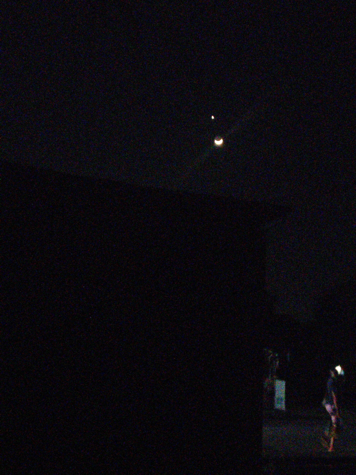 The image captures a dark, nocturnal scene, likely outdoors, with a grainy texture indicative of low-light photography. In the upper portion of the frame, two celestial bodies are visible against the blackness of the sky: a small, bright dot, possibly a star or planet, and a crescent-shaped object, which is likely the moon. The lower portion of the image is dominated by darkness, with faint, blurry shapes suggesting structures or objects. Towards the bottom right, a figure is partially illuminated, appearing to be wearing brightly colored clothing, possibly in the process of walking or standing. Another indistinct figure is visible to the left of the illuminated figure, near a structure that might be a building or stall. The overall impression is of a dimly lit urban or semi-urban environment at night. Without further information or context, pinpointing the exact location within Uyo, Nigeria is not possible from the visual data alone.