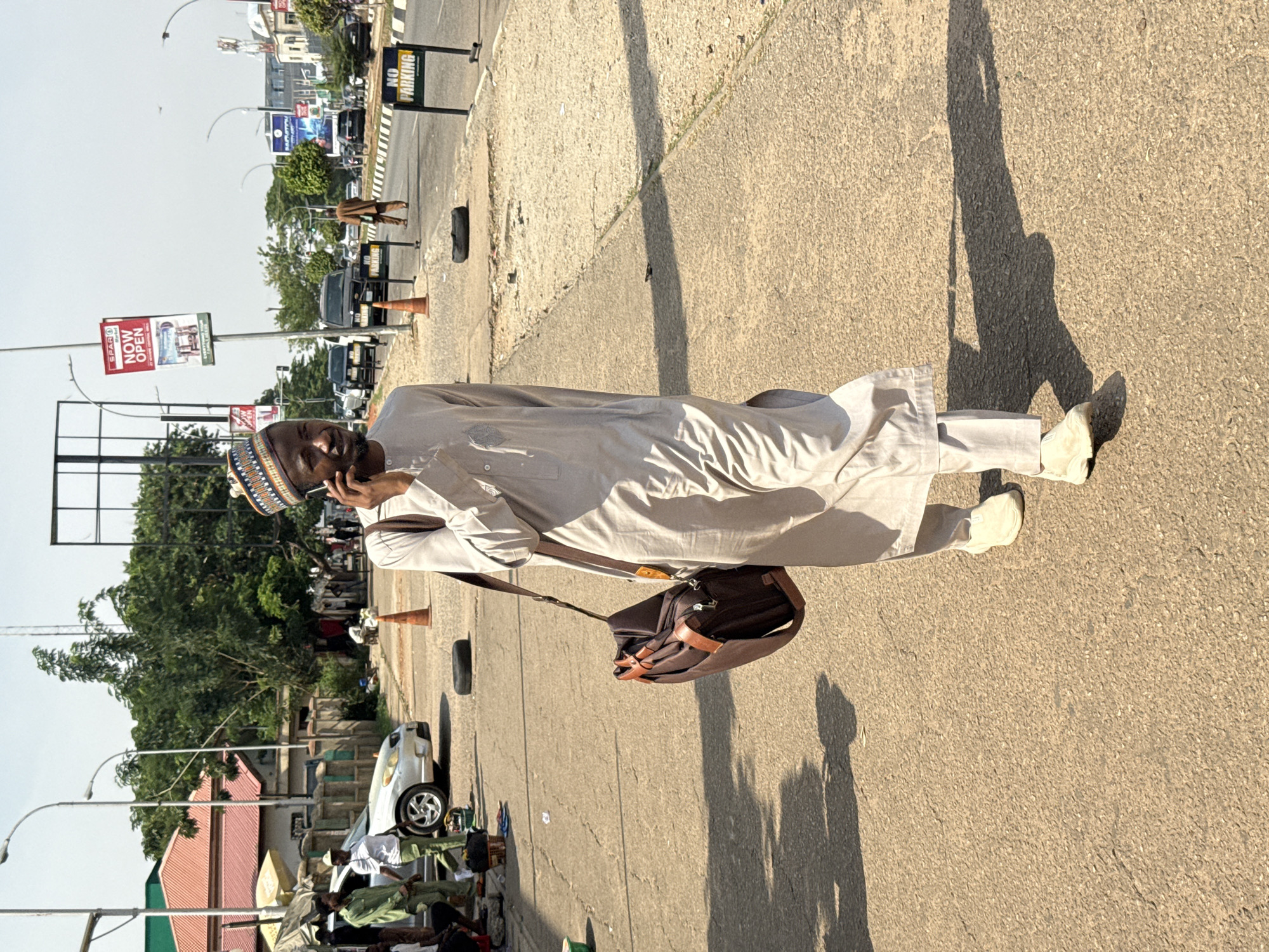 A man in a light-colored traditional Nigerian outfit with a matching cap is standing on a sidewalk in Abuja, Nigeria. He is talking on a cell phone and has a brown messenger bag slung over his shoulder. The sidewalk is cracked and dusty. The shadow of the man stretches out in front of him. In the background, there is a street with cars, buildings, trees, and various street signs. Several figures are seen seated on mats, with their wares for sale displayed in front of them. A white car is parked nearby. A street light is visible, along with a sign that says "NOW OPEN." The sky is clear.