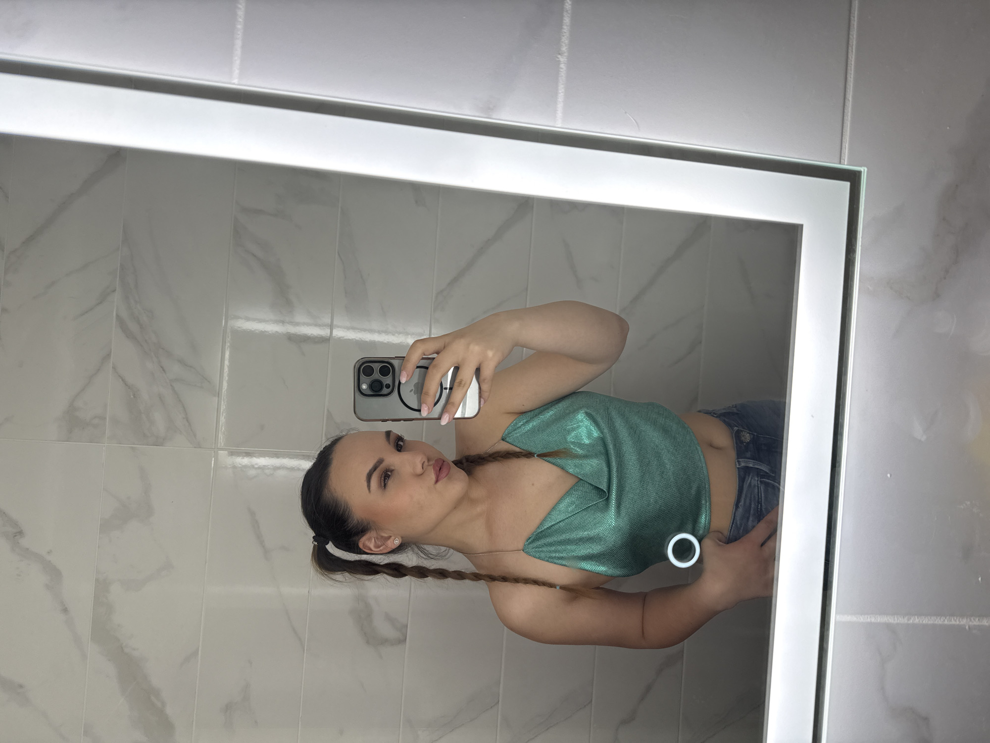 A woman is taking a selfie in a bathroom with white tile walls. She is wearing a green top and blue jeans. She is holding a phone in her right hand and posing for the camera. She has brown hair styled in pigtails. The mirror has a built-in light around the edge. The location is not possible to determine from the image, so Sofia, Bulgaria cannot be identified.